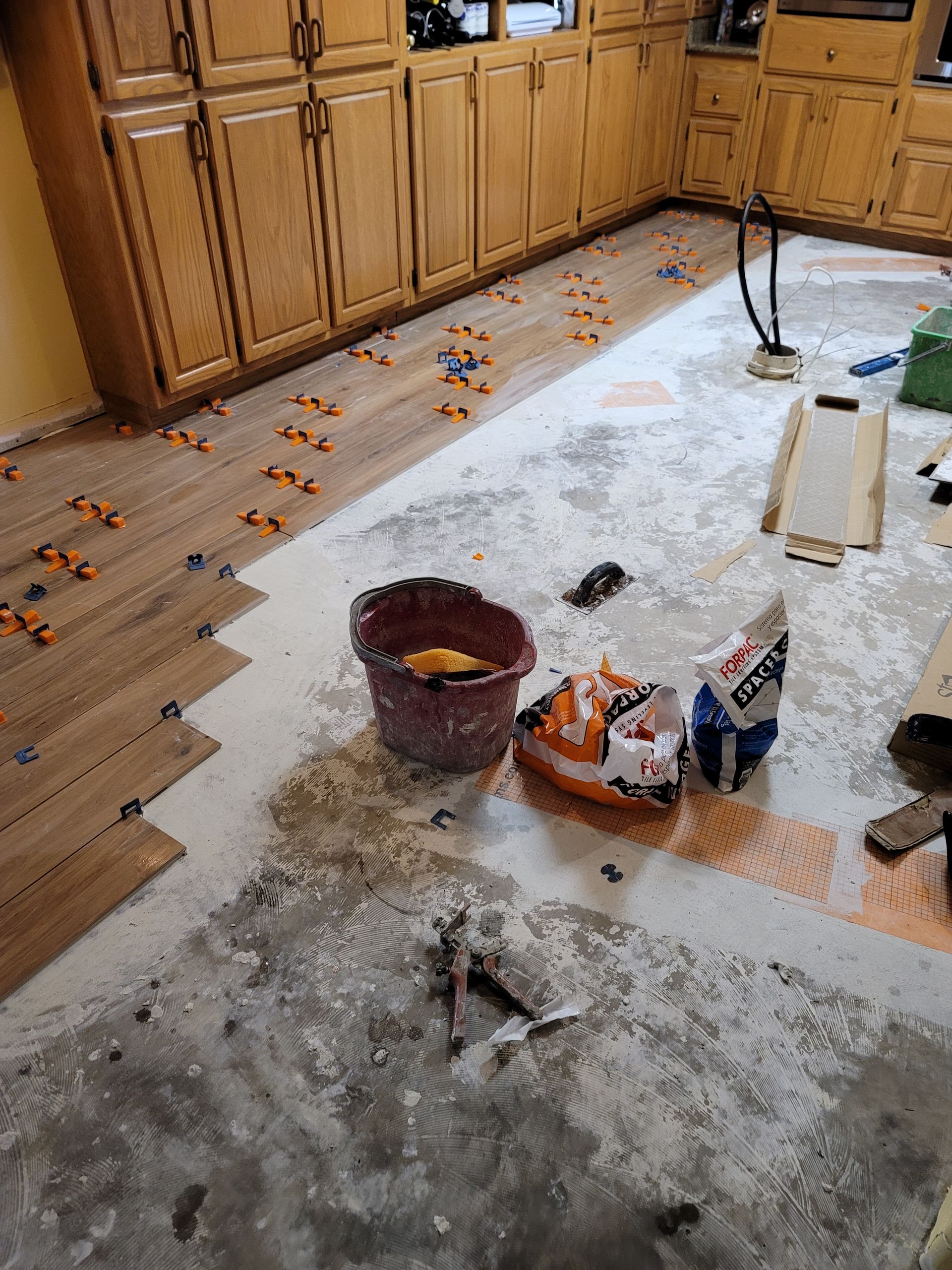 Messy kitchen floor with spilled debris, bucket, and cleaning supplies near wooden cabinets