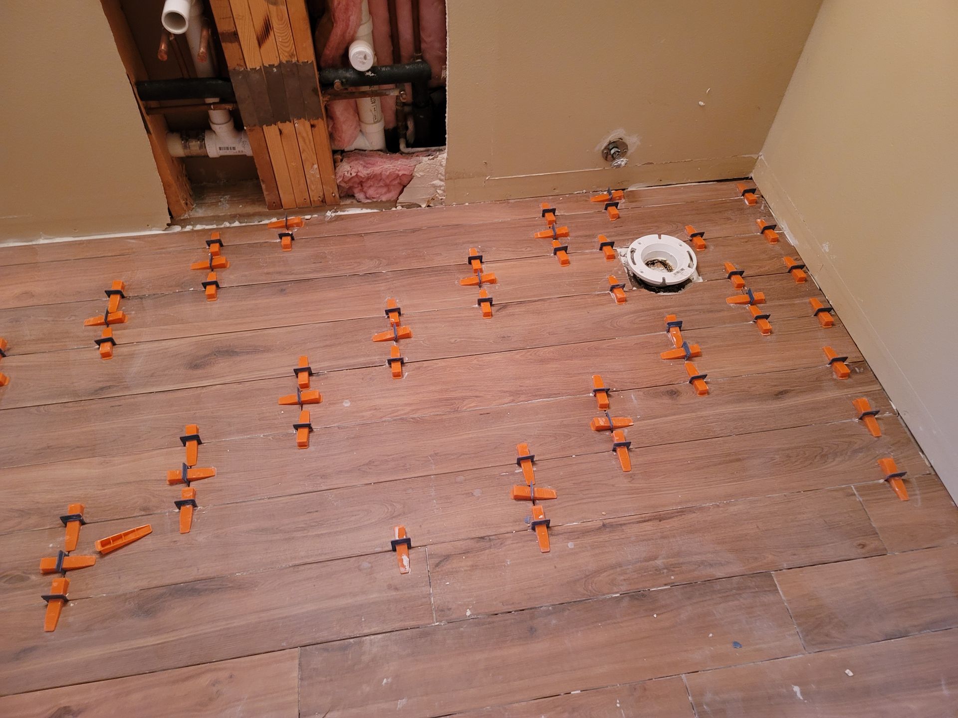 Flooring under renovation with orange leveling clips and a white drain opening near a cutout wall opening