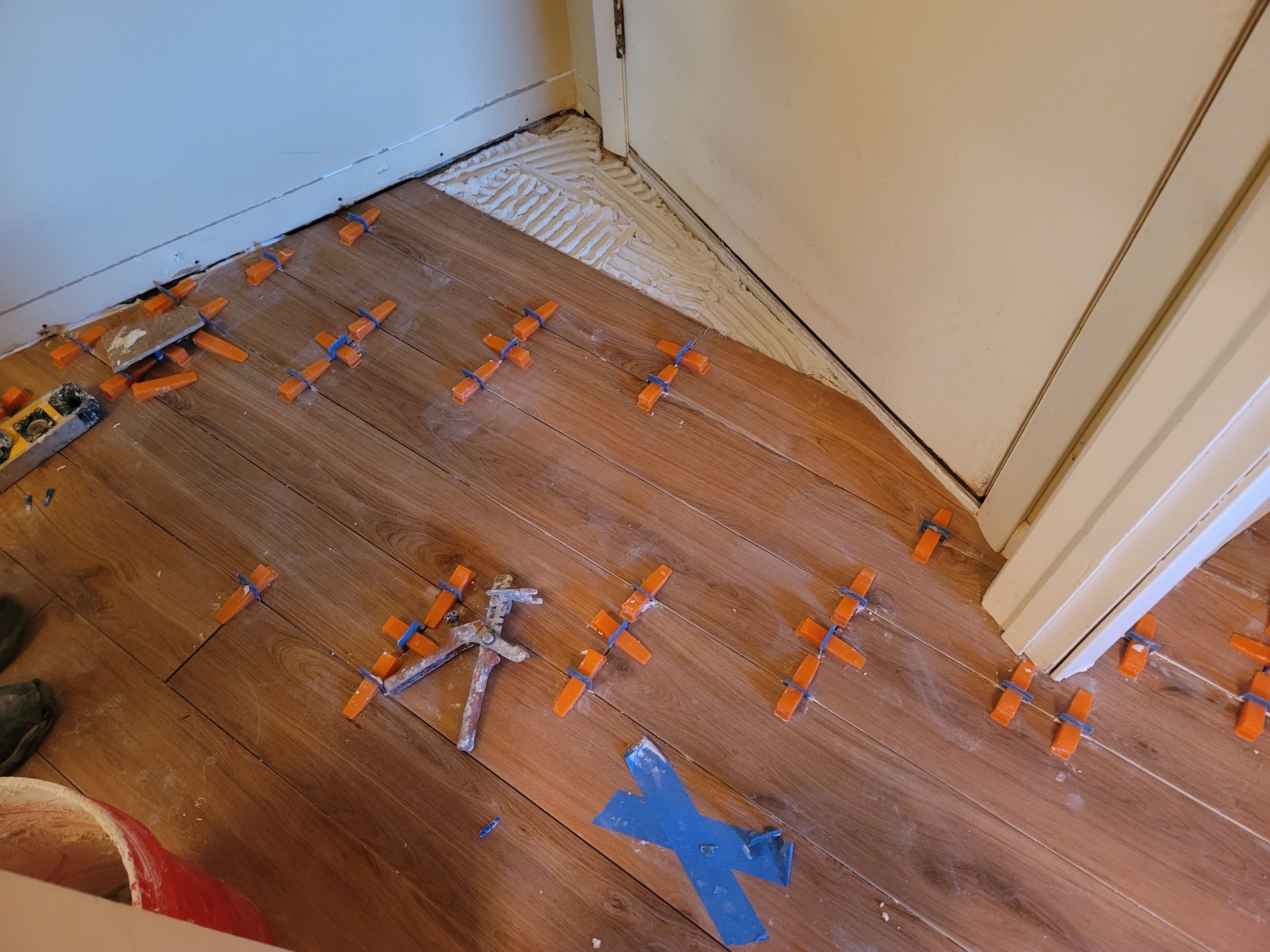 Partially installed wood floor with orange adhesive strips and a blue tape marking near a corner wall.