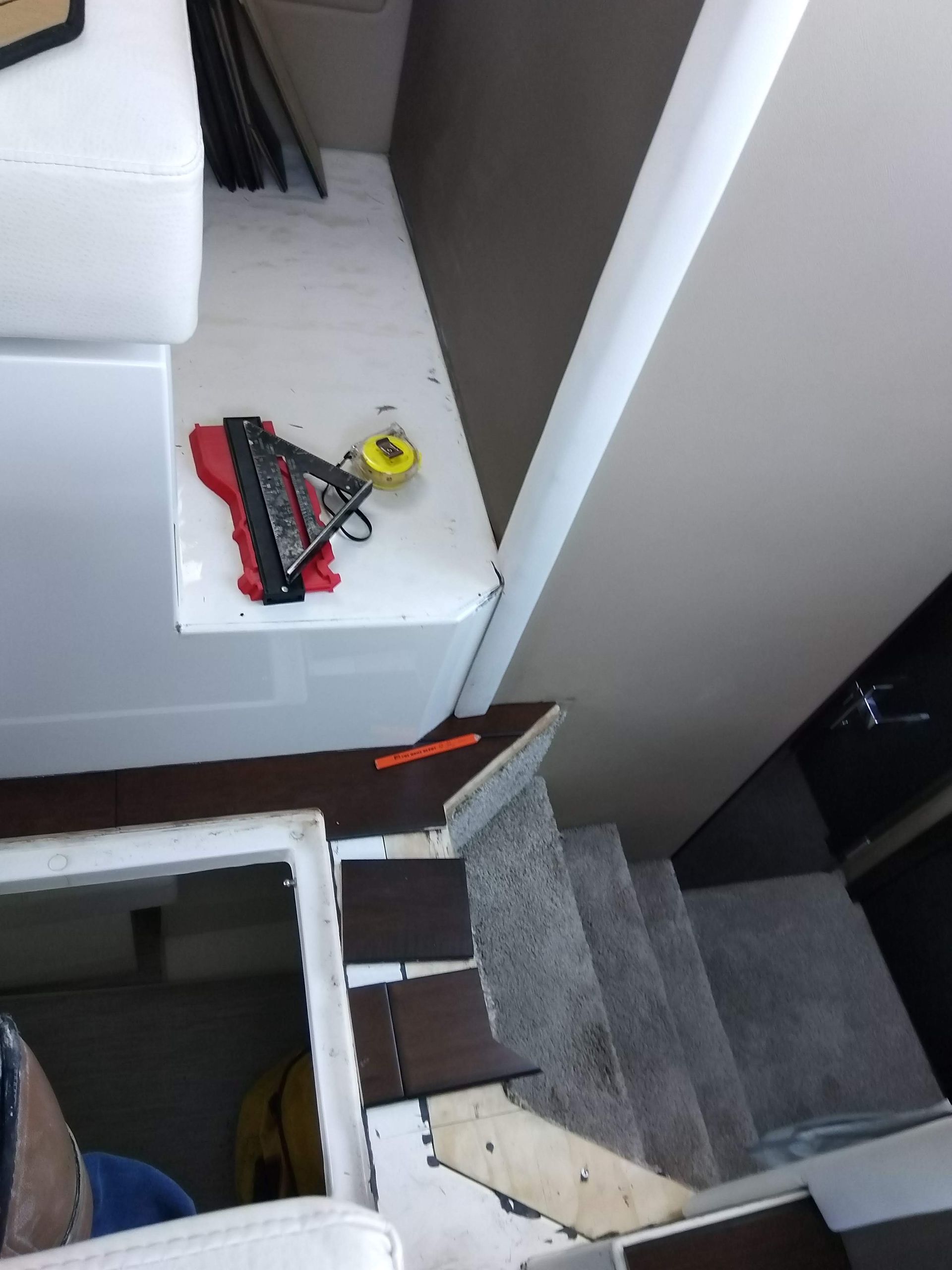 Stairs with gray carpet; white landing holds a red-and-black tool and yellow tape measure near an open stairwell.