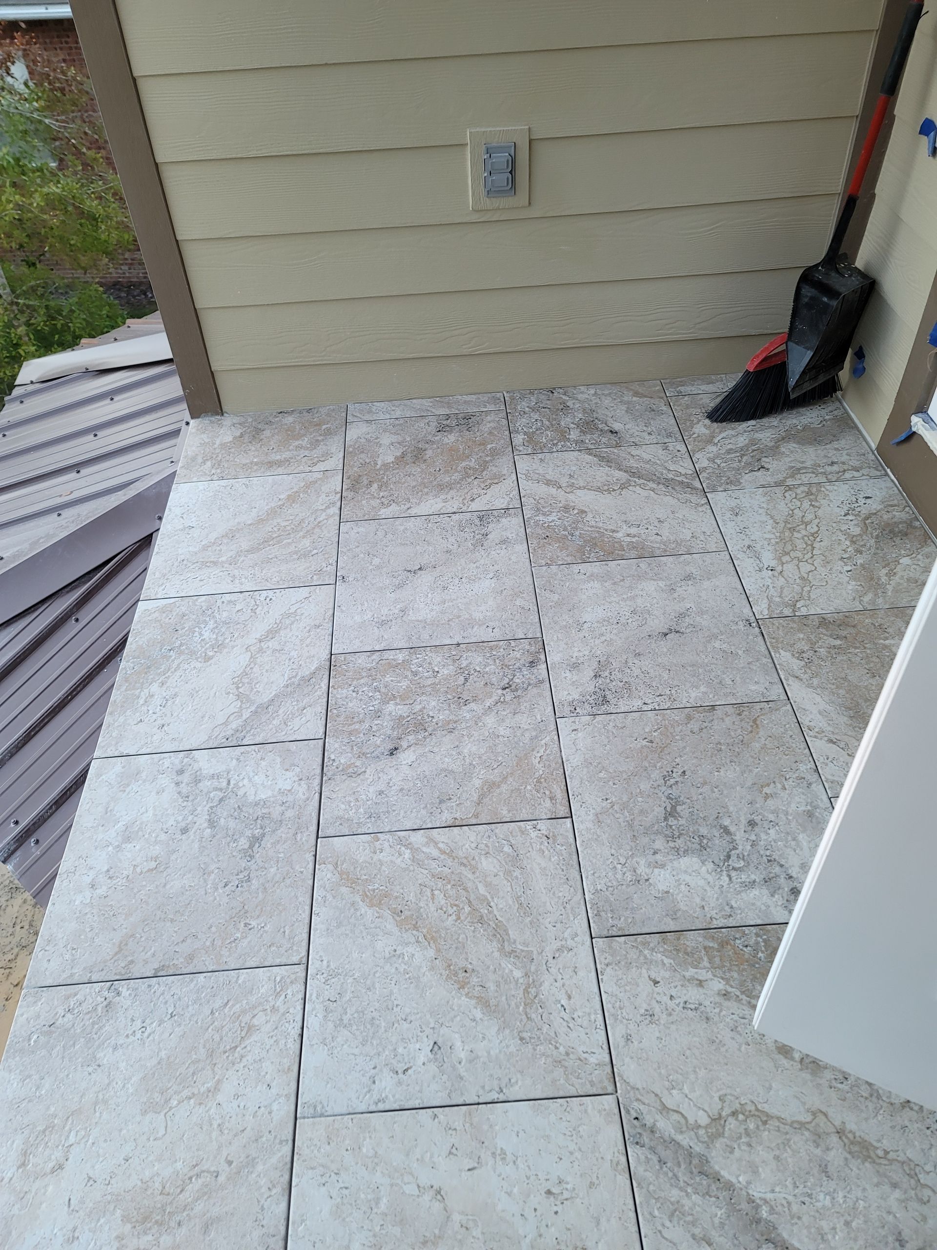 Outdoor patio with large pale stone tiles beside a beige house wall and a red-handled shovel.