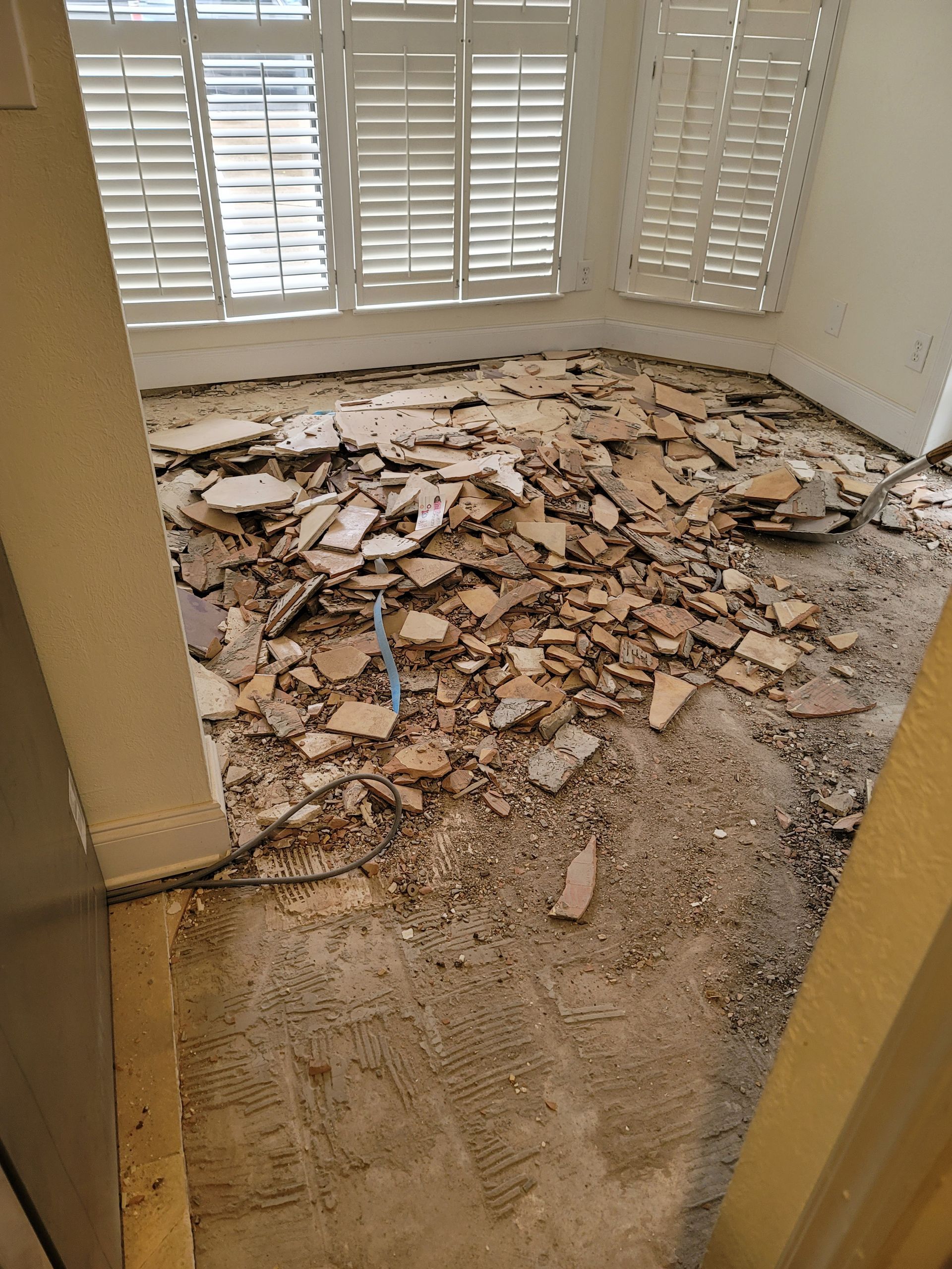 Floor covered with broken tiles and debris in a sunlit room with white shutters
