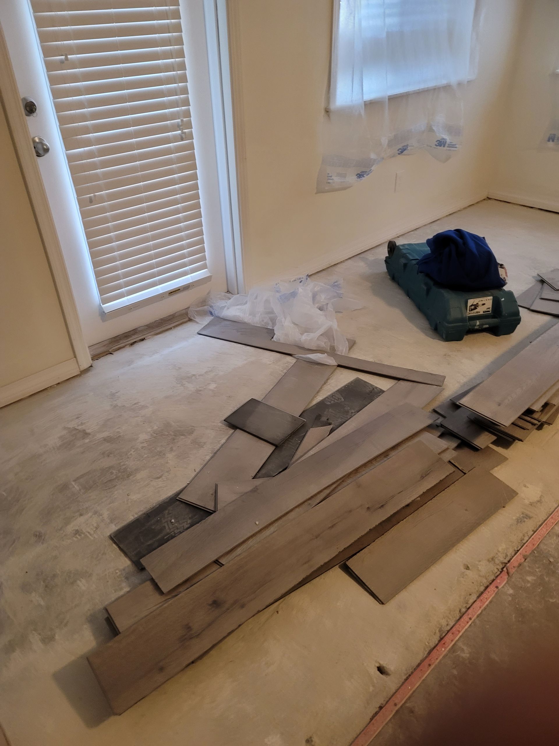 Room with white walls and a wood floor being installed, with scattered planks and tools near a door and window.