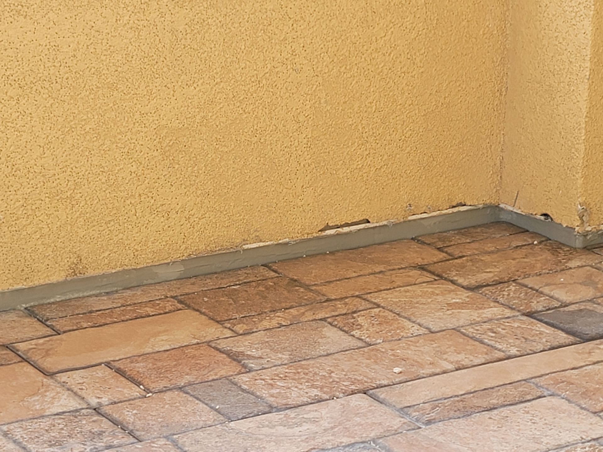 Empty corner with yellow walls and a tiled floor, showing a gray baseboard along the wall.