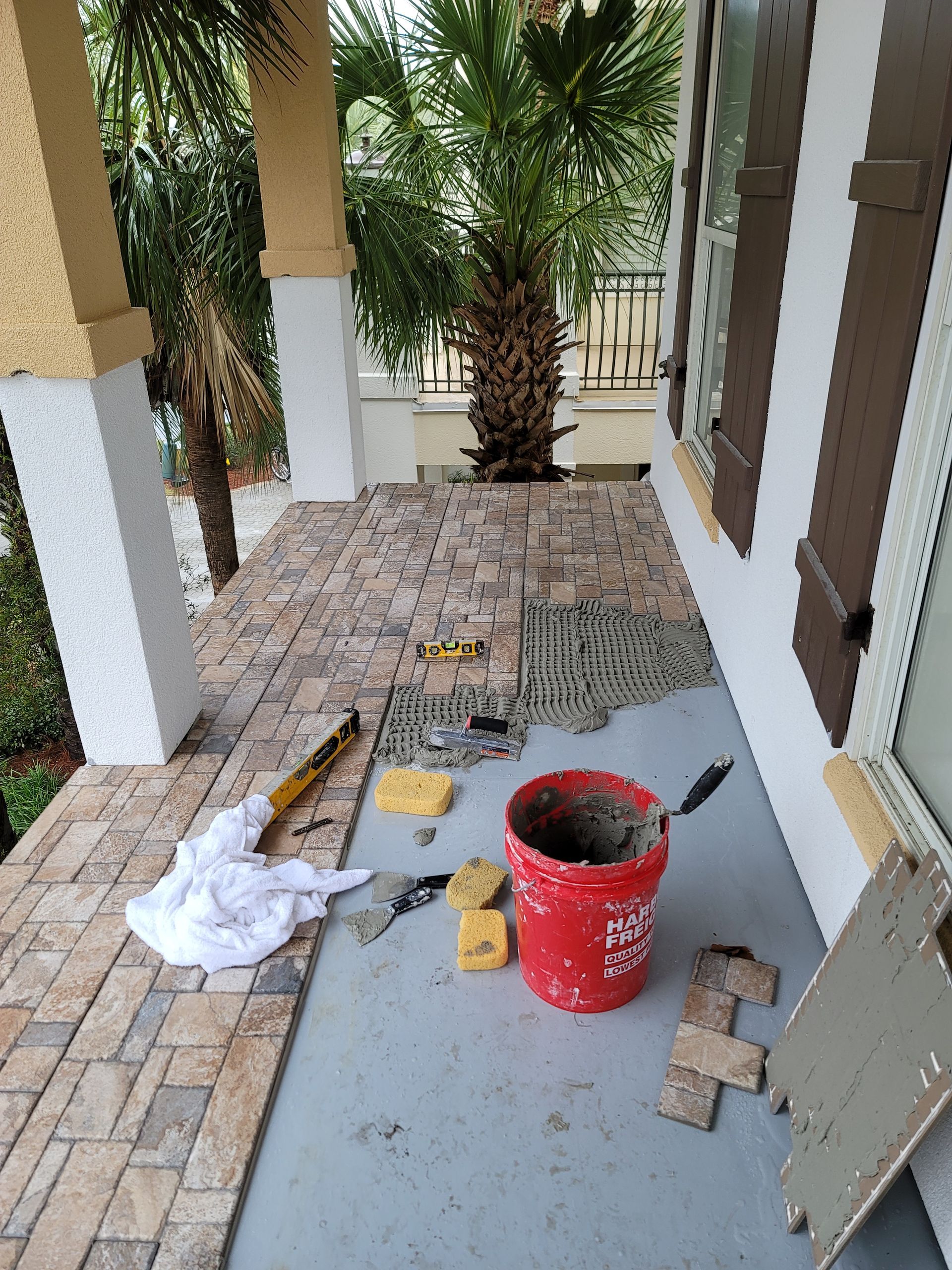 Patio under construction with scattered pavers, tools, and a red bucket beside porch columns
