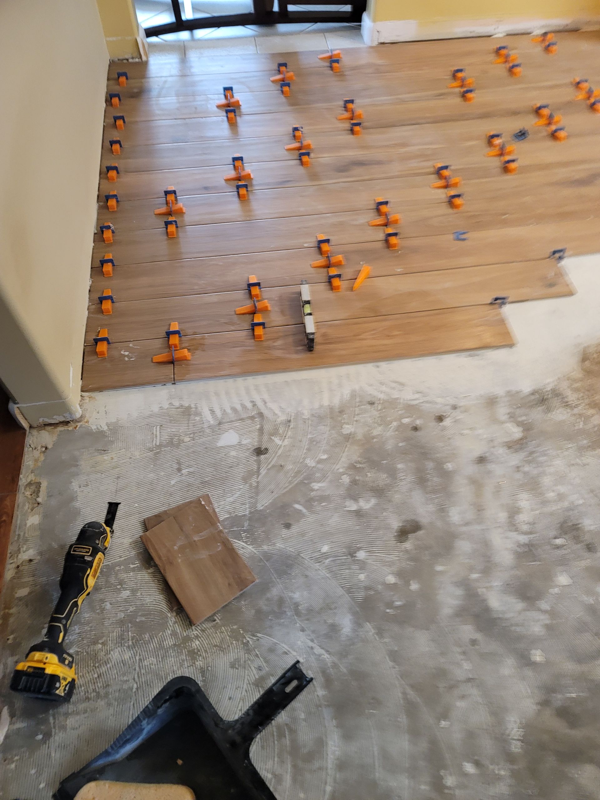 Flooring installation in progress with wood planks, orange spacers, and a drill on a concrete subfloor.