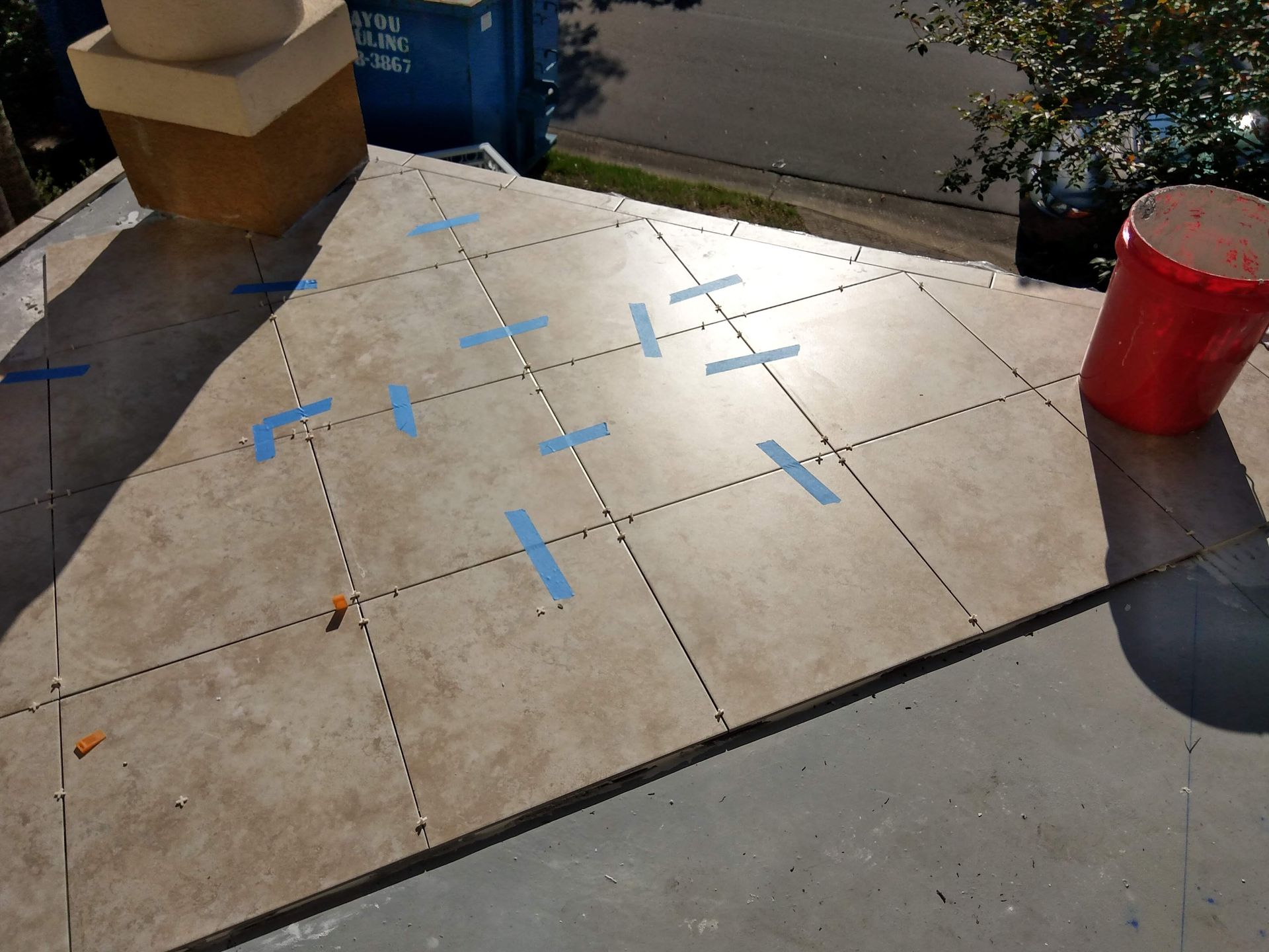 Outdoor tiled sidewalk marked with blue tape, beside a red trash can and a planter box