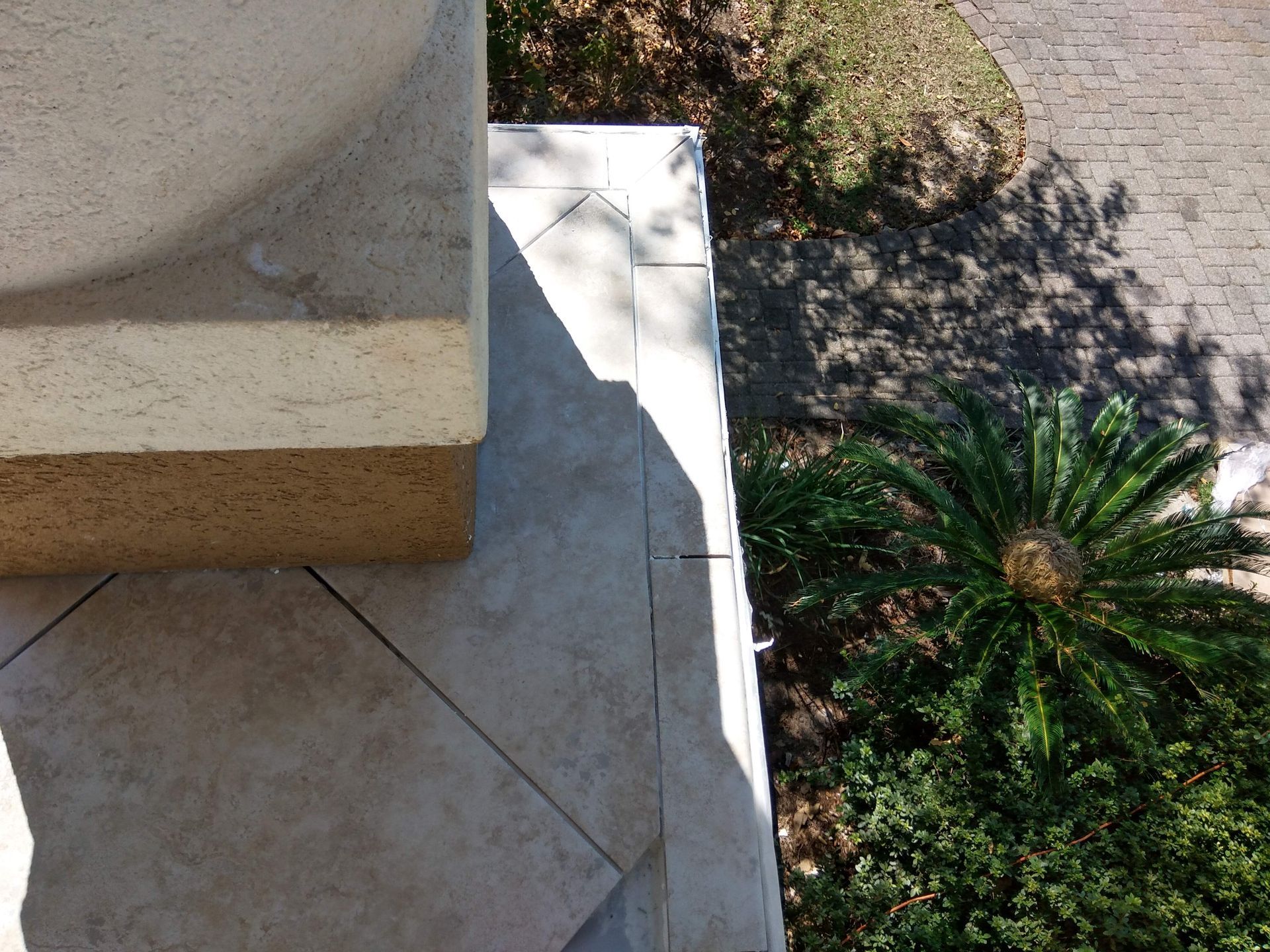 Corner of a tan stucco building beside a tiled patio and a shaded garden path with a palm shrub.
