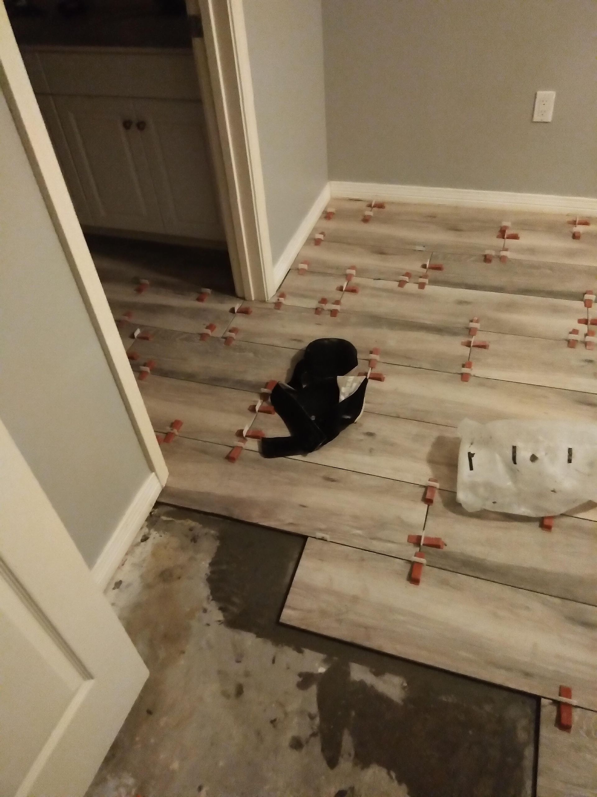 Partially laid flooring in a room, with a black cat sitting near the center and tools on the floor.