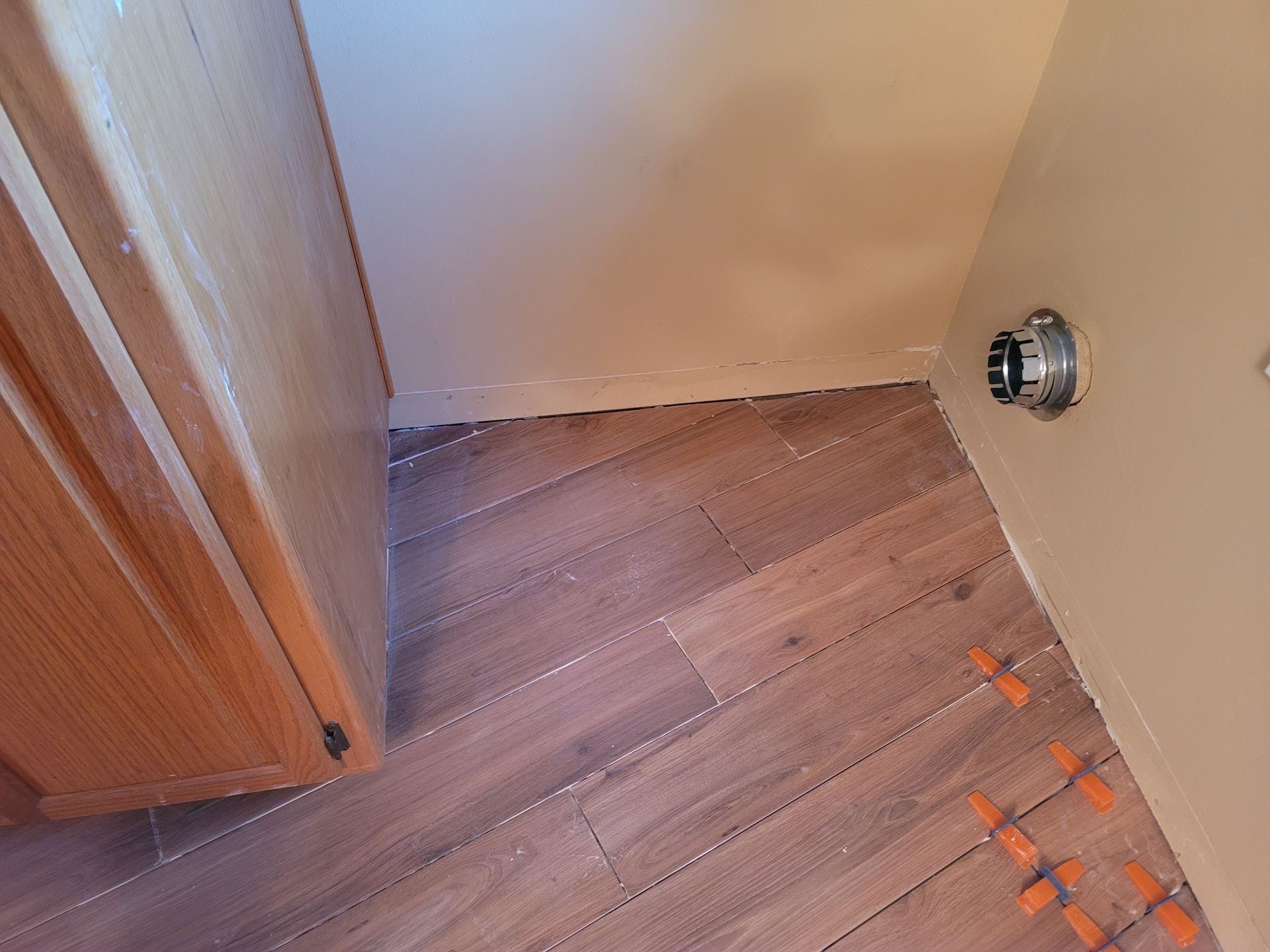 Small bathroom corner with wood floor, beige walls, cabinet edge, and orange tile spacers near a shower fixture.