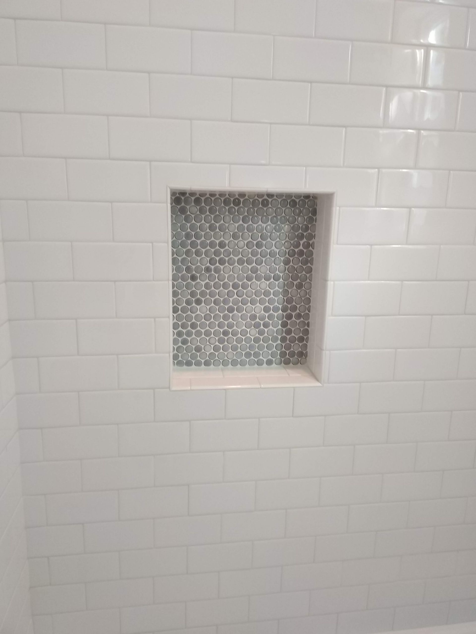White tiled shower wall with a recessed niche lined in gray patterned mosaic tile.