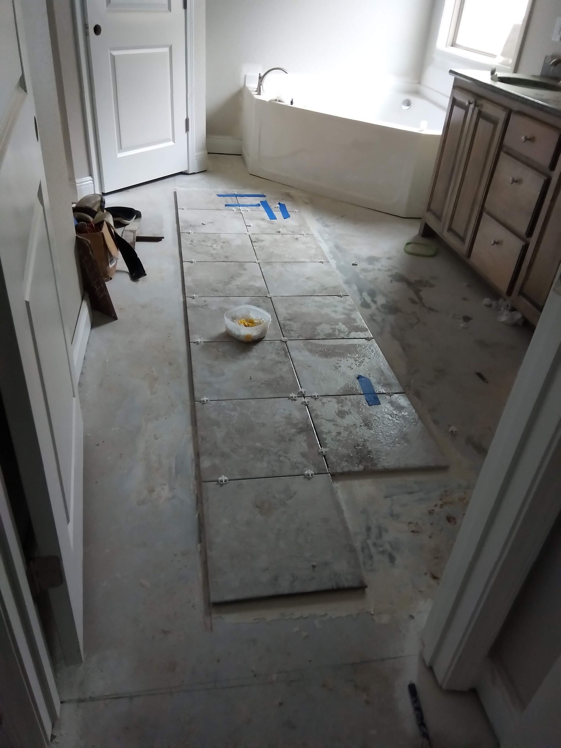 Bathroom under renovation with floor tiles laid out and tools near a tub and vanity.