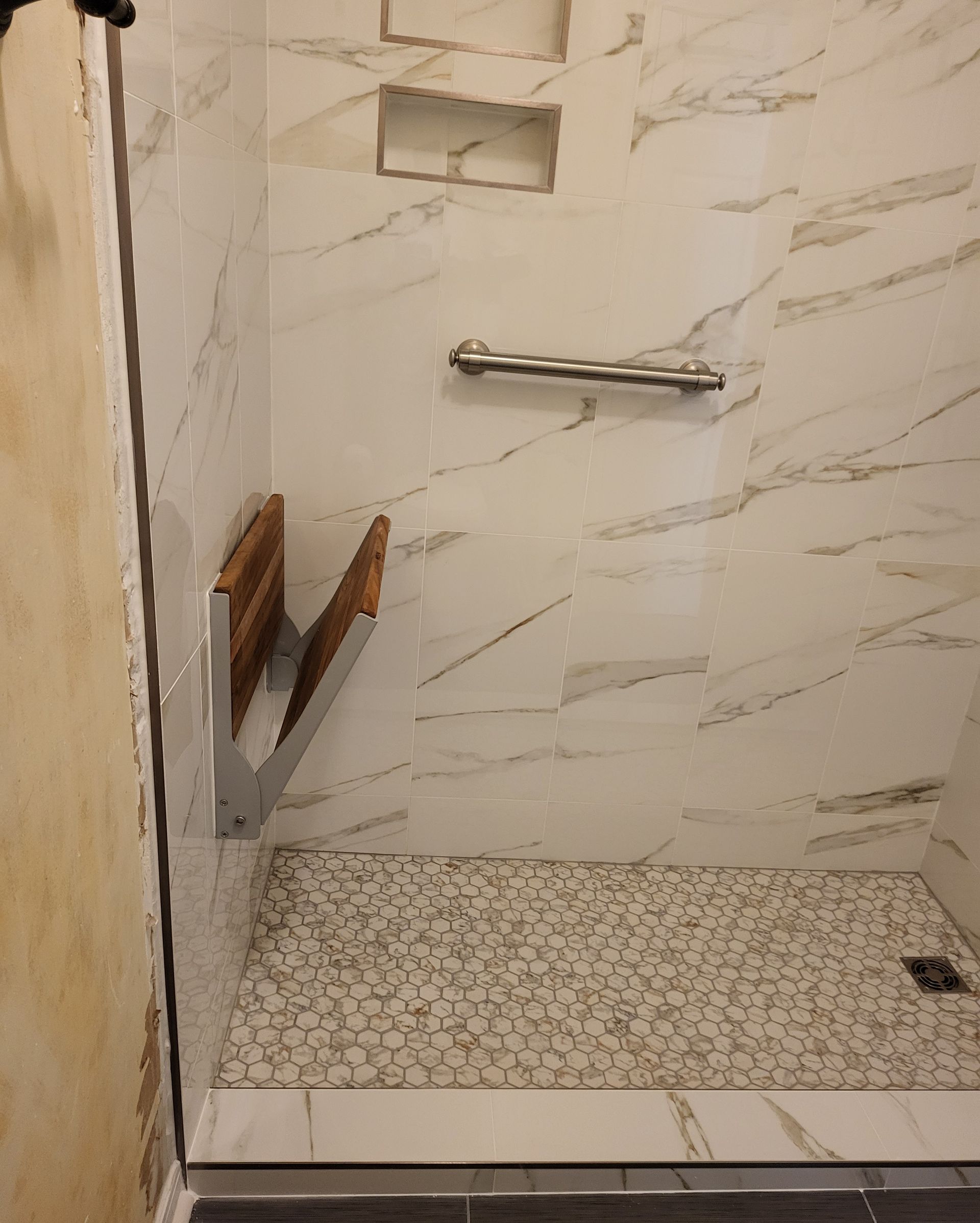 Small tiled shower stall with marble-look walls, a grab bar, and a soap shelf.