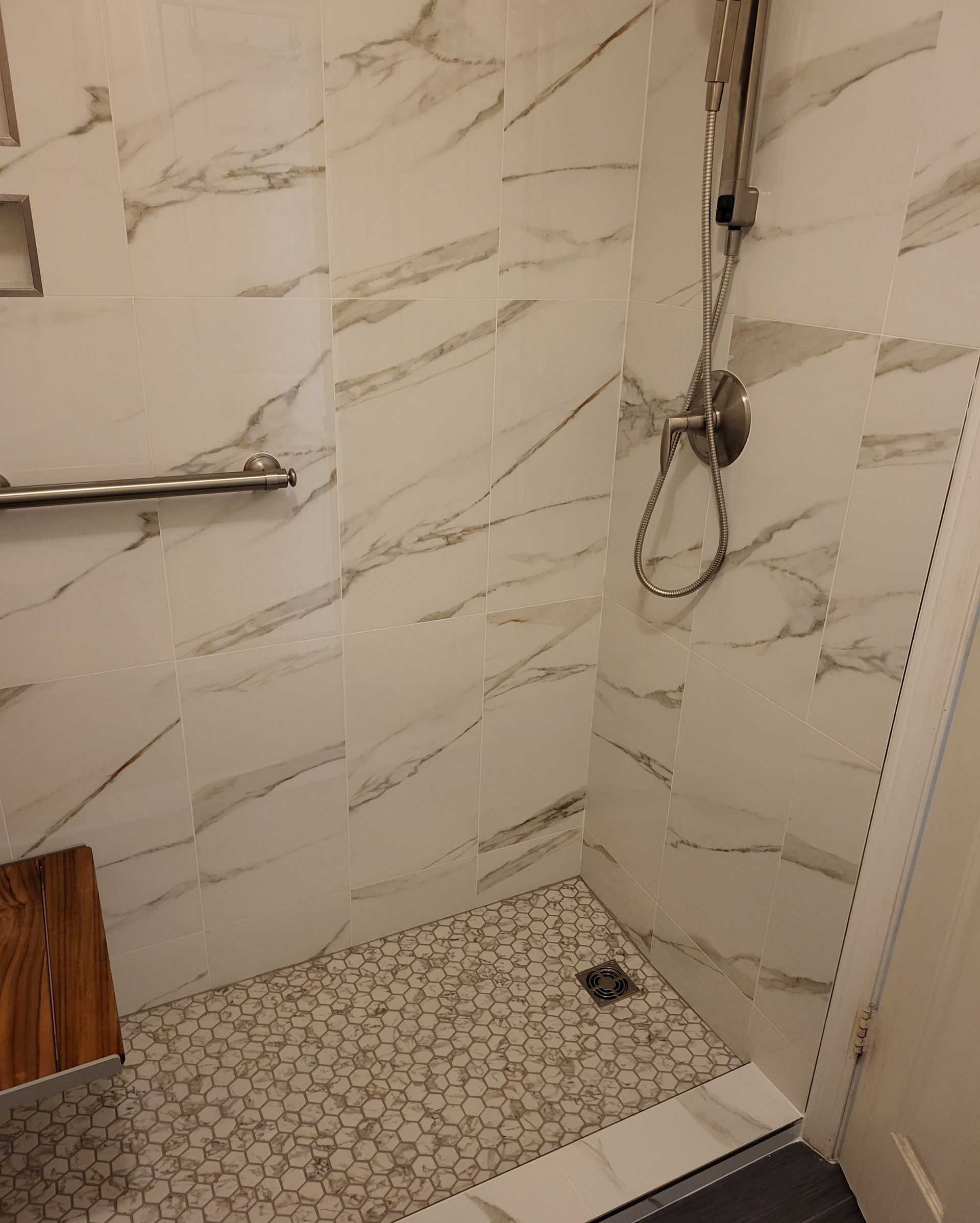 Small tiled shower stall with chrome grab bar, handheld showerhead, and patterned shower floor drain.