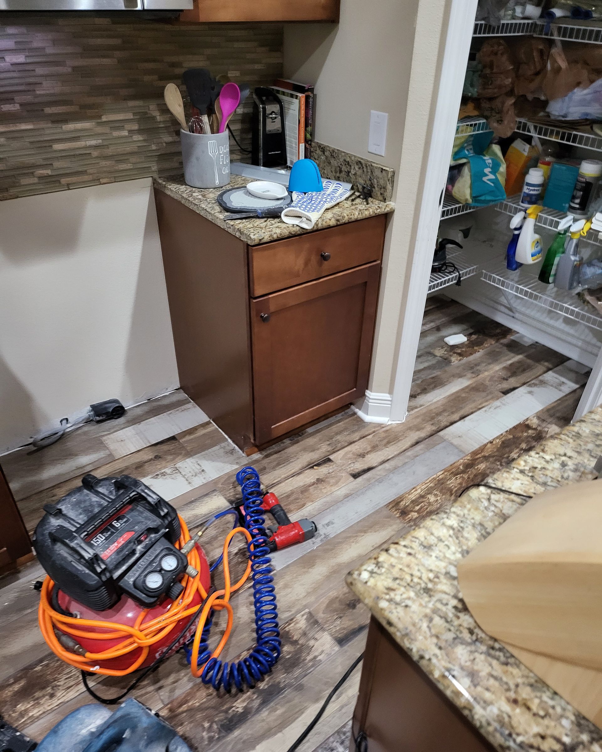 Kitchen floor with power tools, coiled cords, and a blue hose beside an open pantry and cabinet.