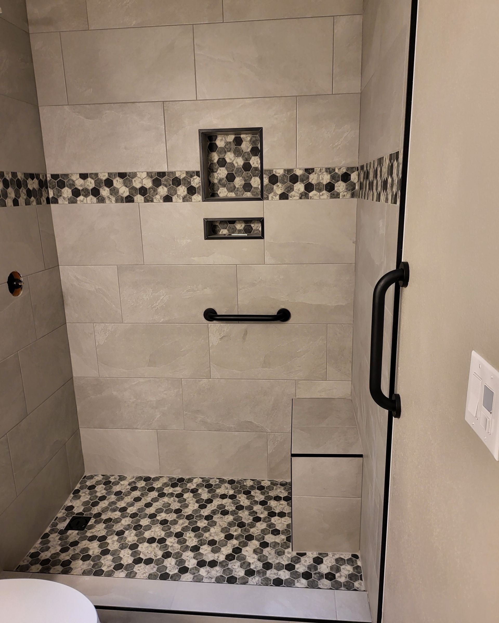 Accessible shower with grab bars, mosaic tile accents, and a bench seat in a tiled bathroom.
