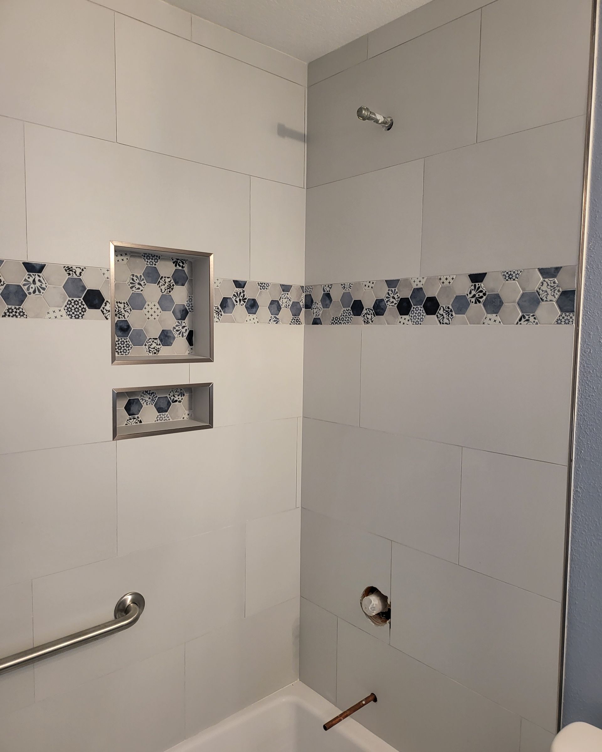Partially tiled bathroom shower with blue floral border, shelves, grab bar, and exposed plumbing holes
