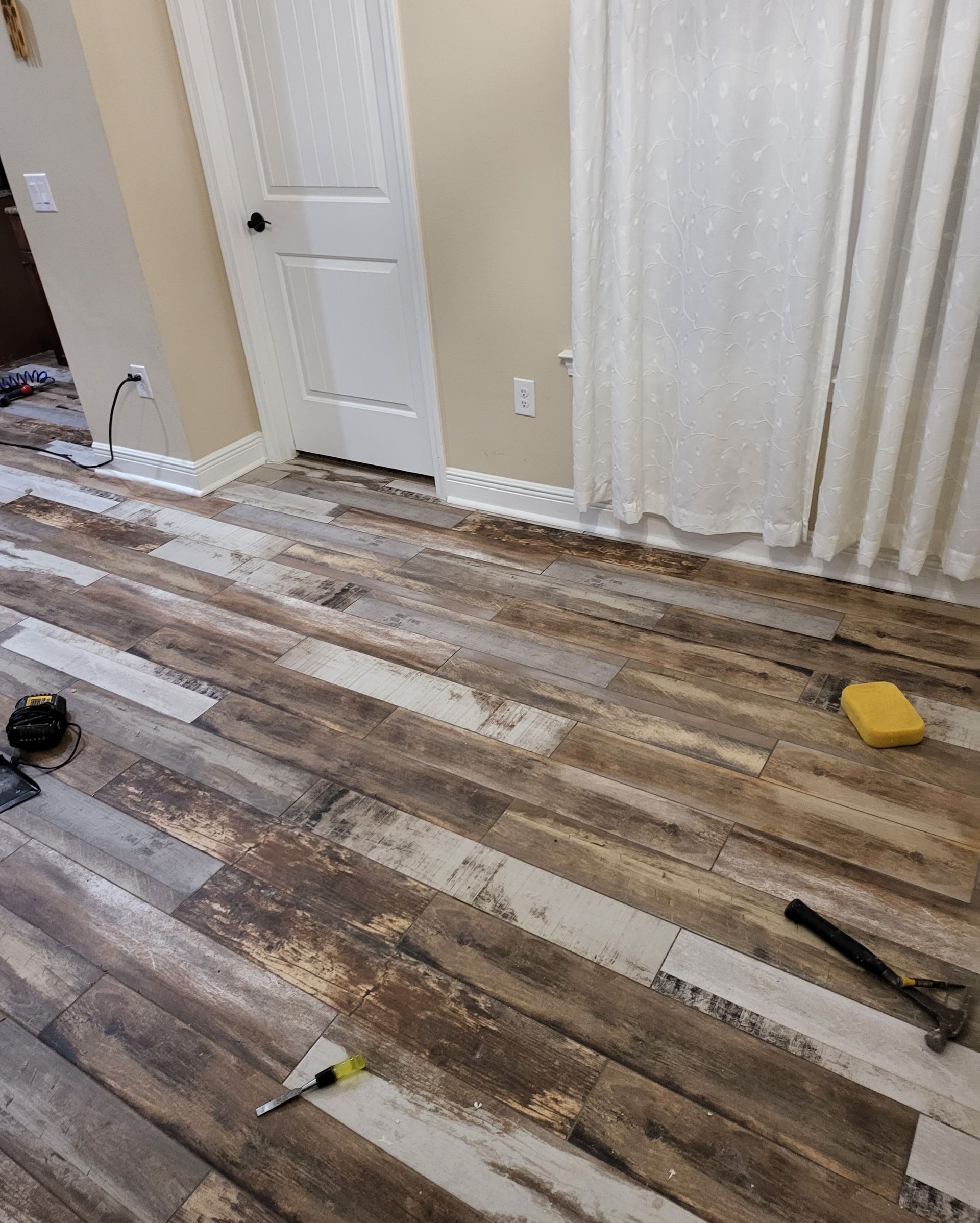 Scattered wood flooring planks in a room with curtains and a white door, tools on the floor.