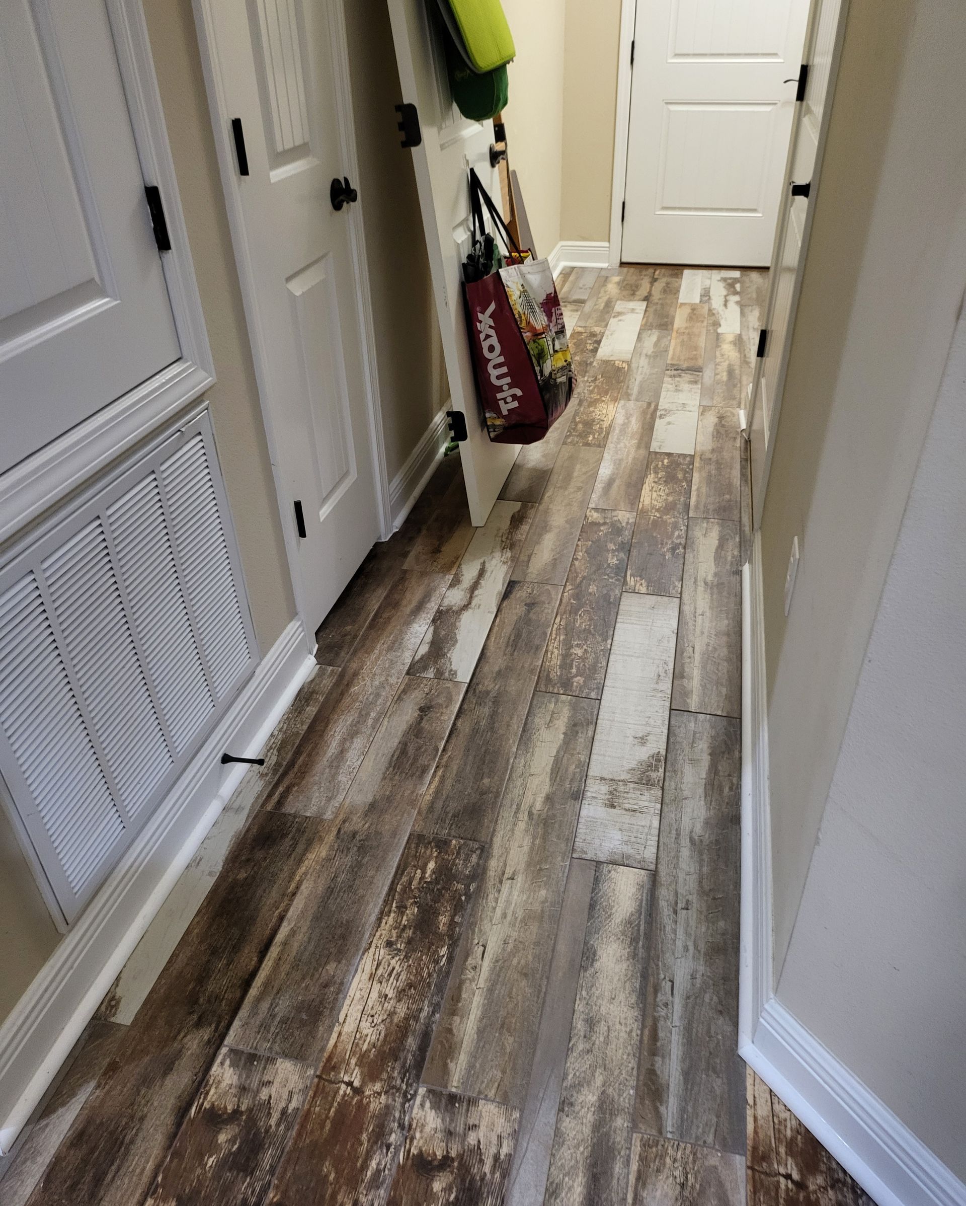 Narrow hallway with gray-brown wood-patterned floor and white doors, a vacuum and mop leaning against the wall.