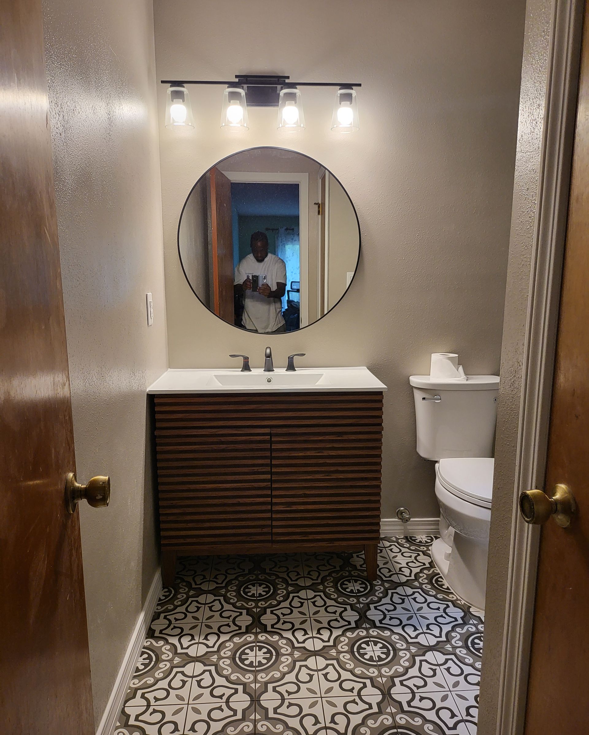 Small bathroom with patterned tile floor, wood vanity, round mirror, and toilet; person reflected in mirror.