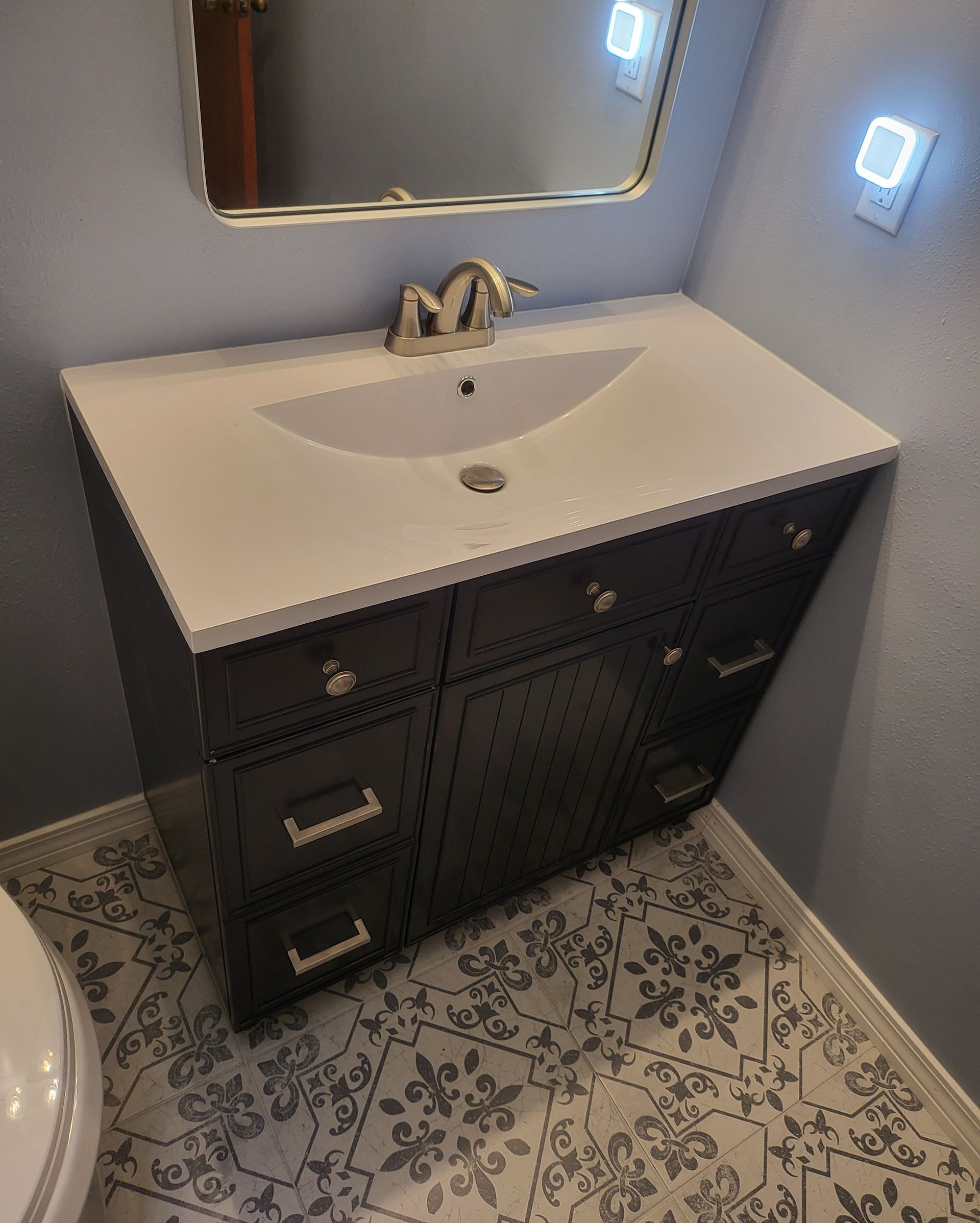Dark vanity with white sink, mirror, and patterned tile floor in a small bathroom.