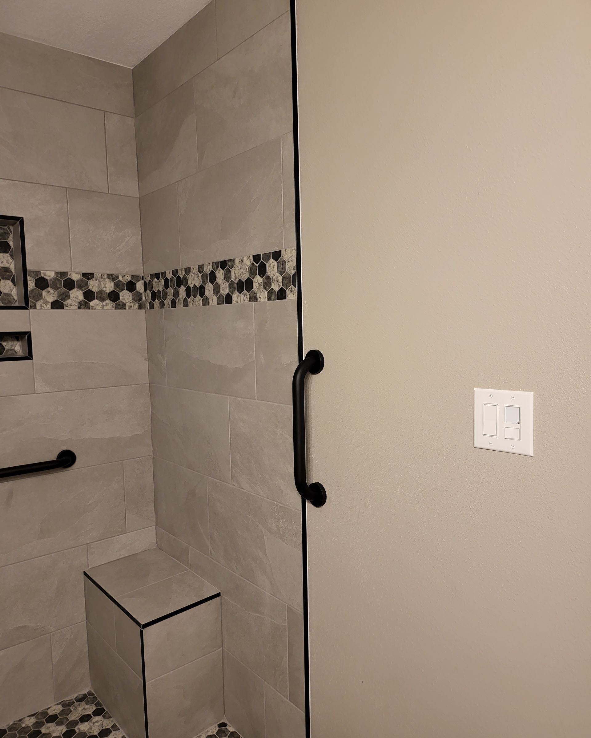 Bathroom shower with gray tile, black grab bars, and a small built-in bench next to a beige wall.