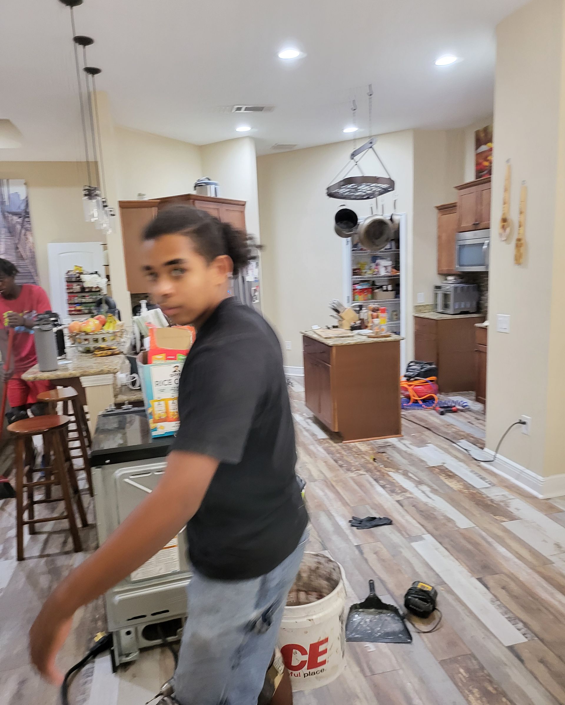 Person in a cluttered kitchen, turning toward the camera with tools and household items on the floor.