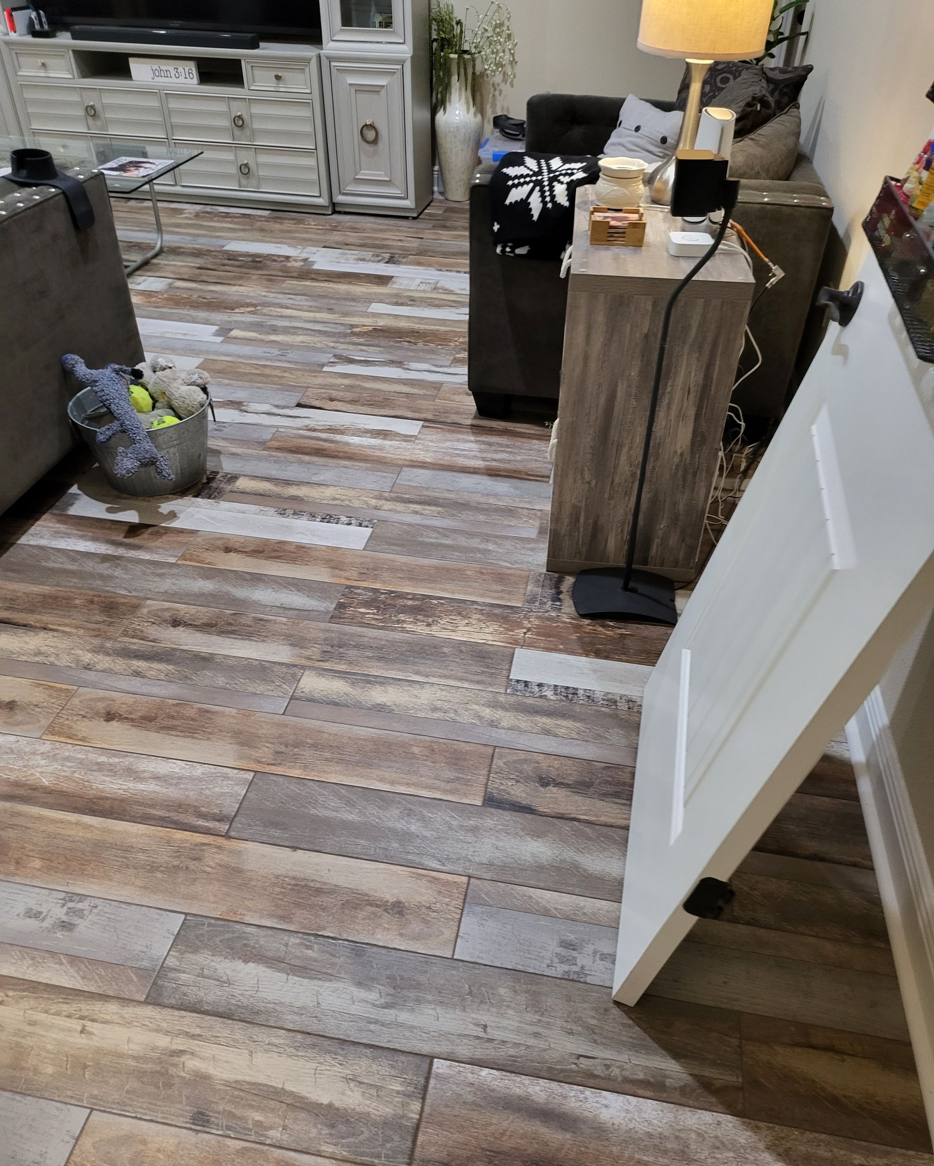 Cluttered living room with wood-look floor, lamp on a side table, and a white door ajar on the right.