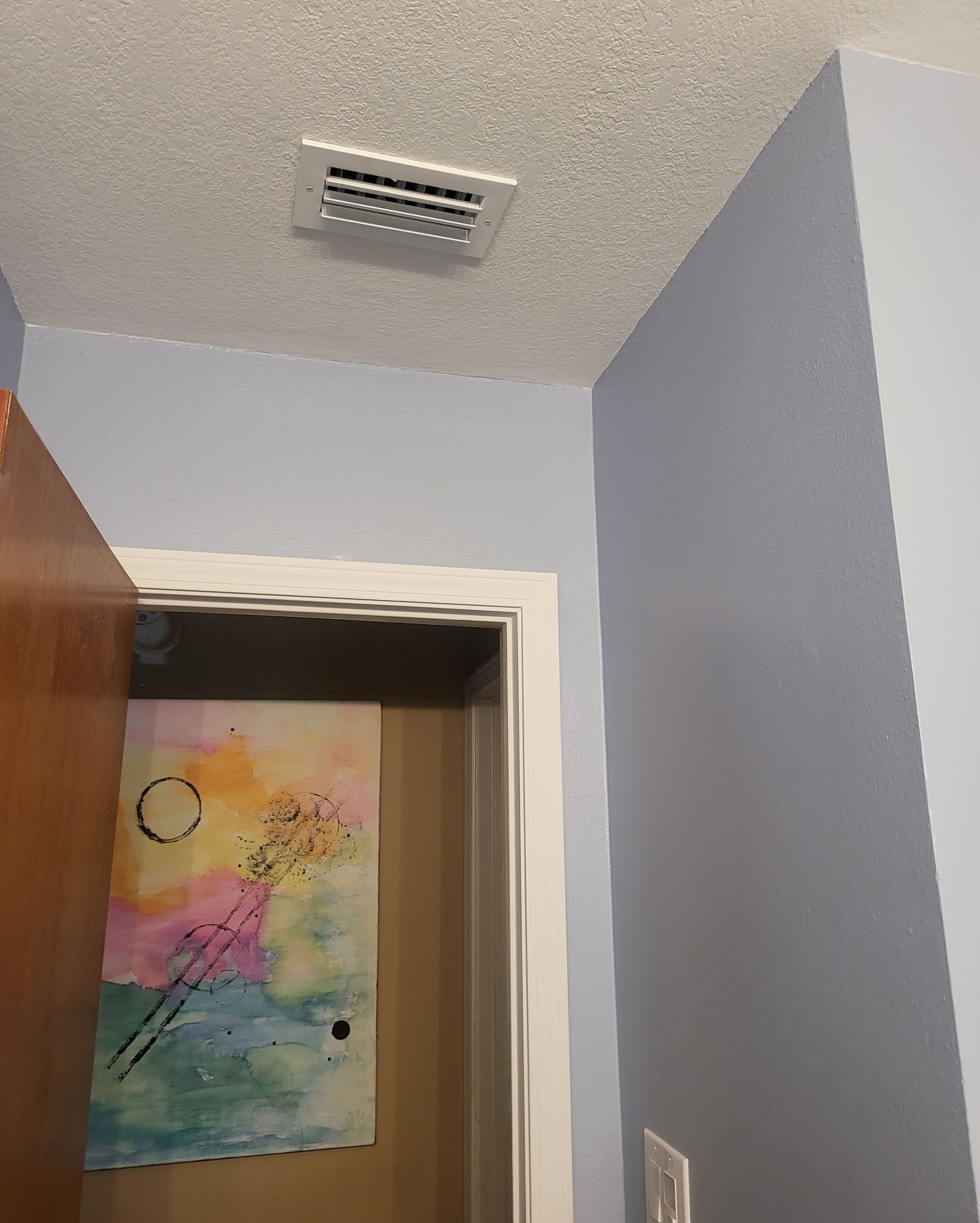 Small room corner with a ceiling vent, light blue walls, and a colorful drawing taped on a doorway.