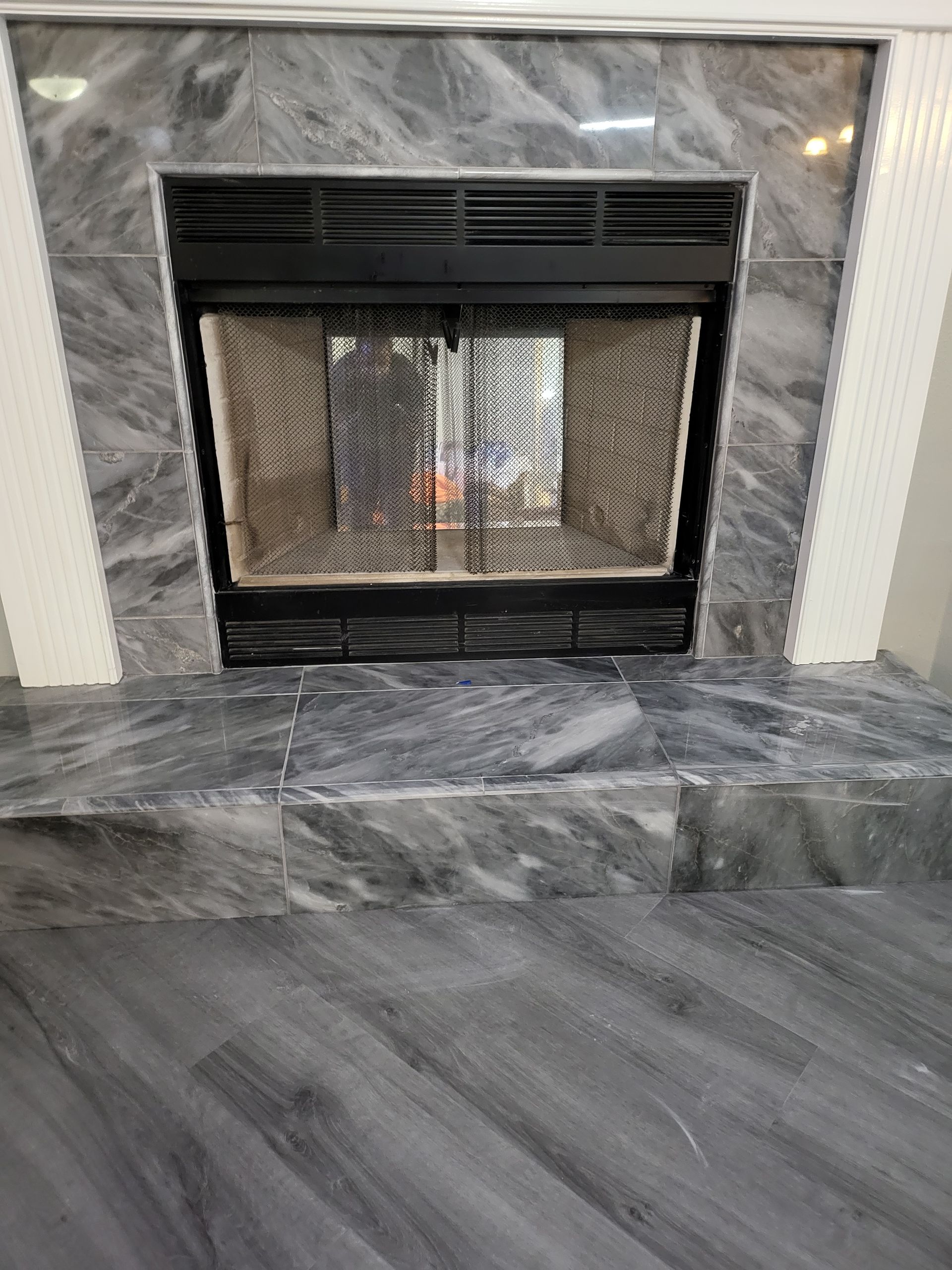 Black fireplace with glass doors and gray marble surround and hearth in a room with gray wood flooring