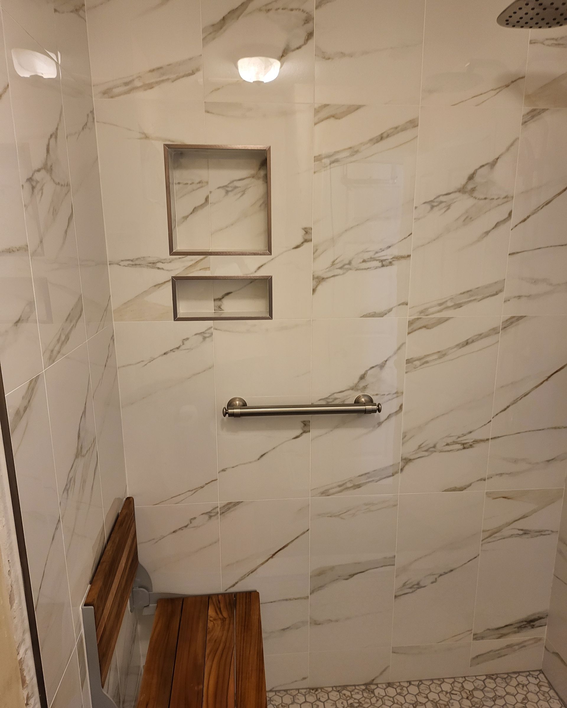 Small tiled shower with built-in shelves, grab bar, and wooden bench seat.