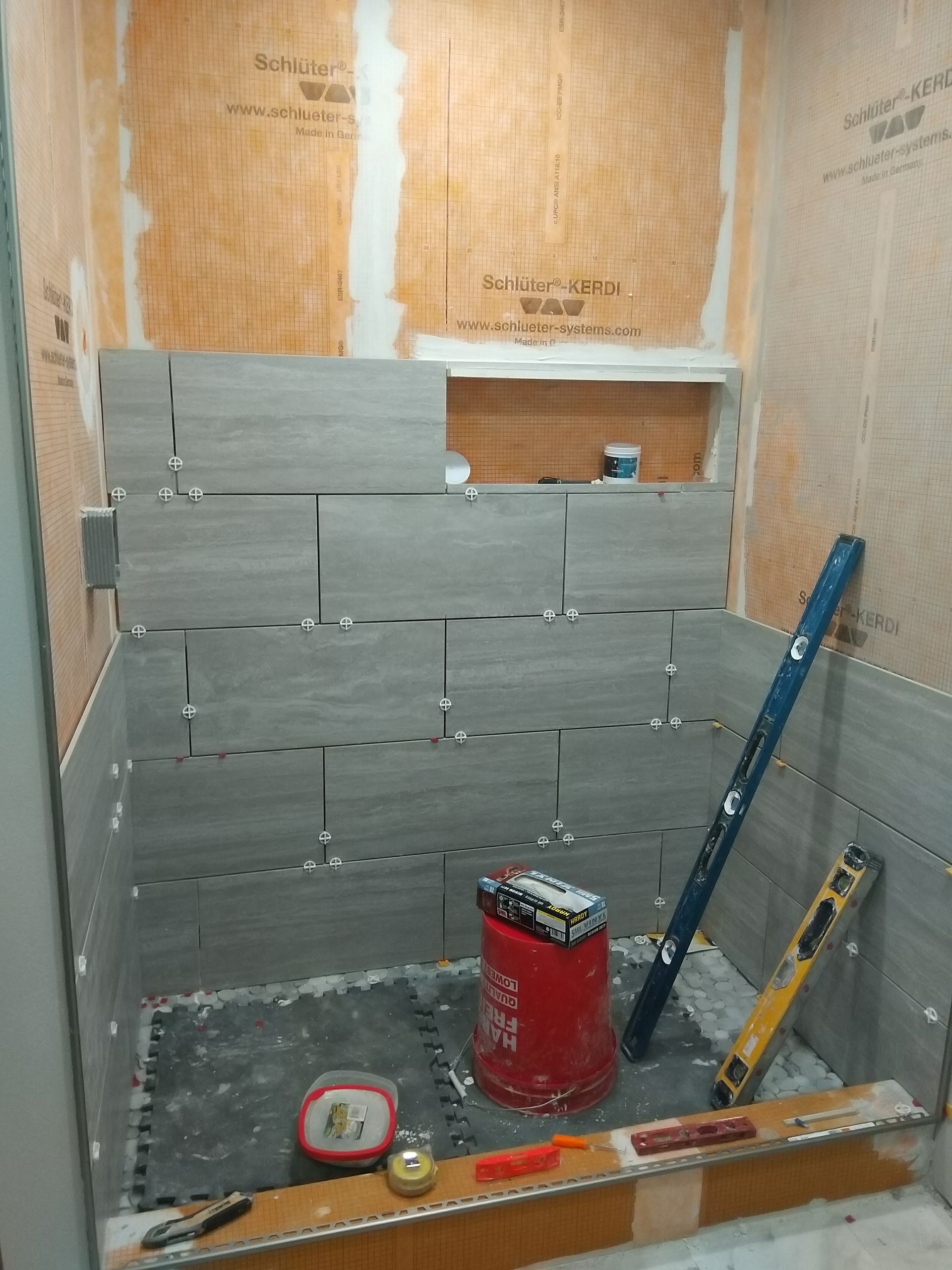Partially tiled wall in a small bathroom under construction, with tools and a red bucket on the floor