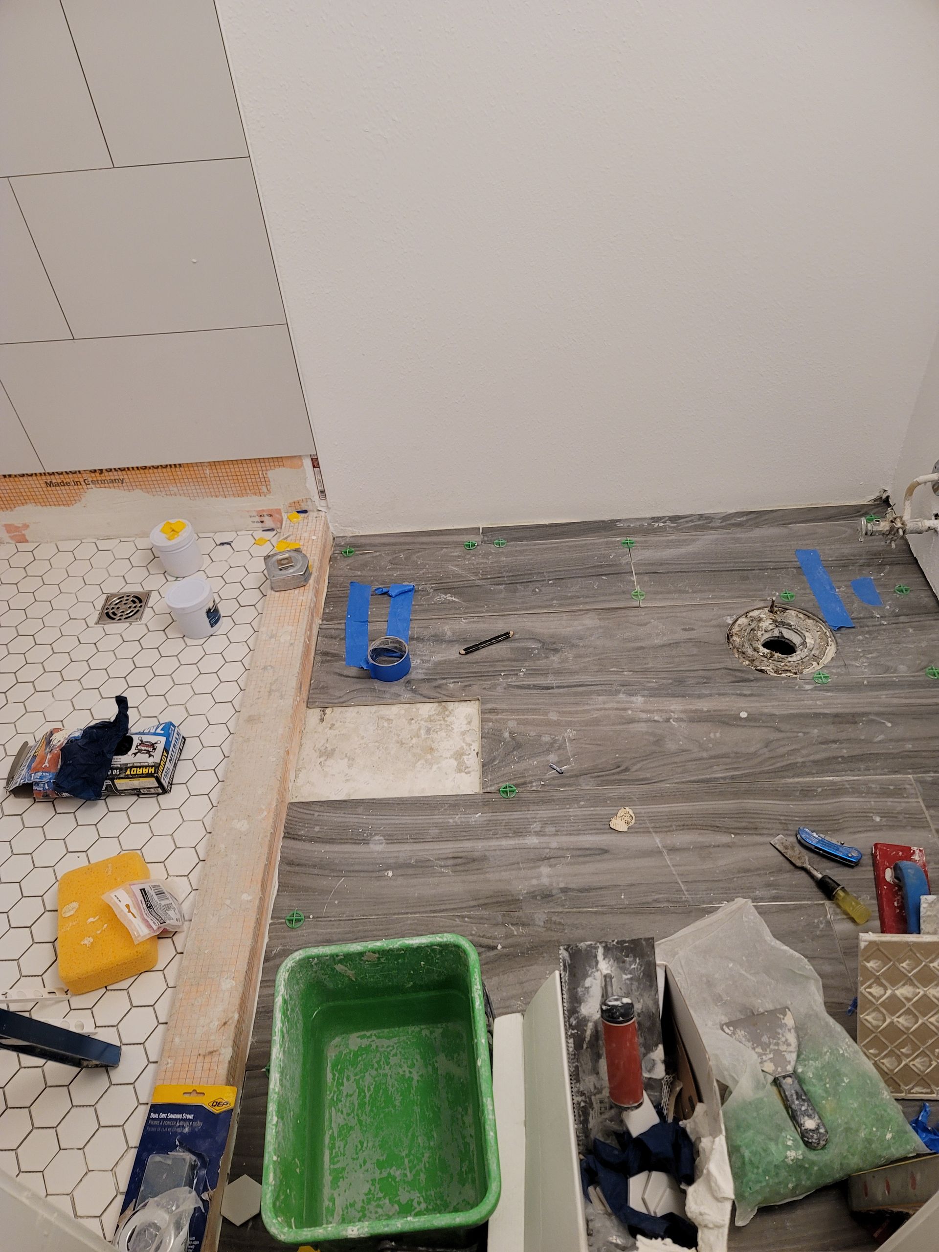 Tool-filled room with unfinished floor, white wall, green bucket, and scattered tiles and supplies