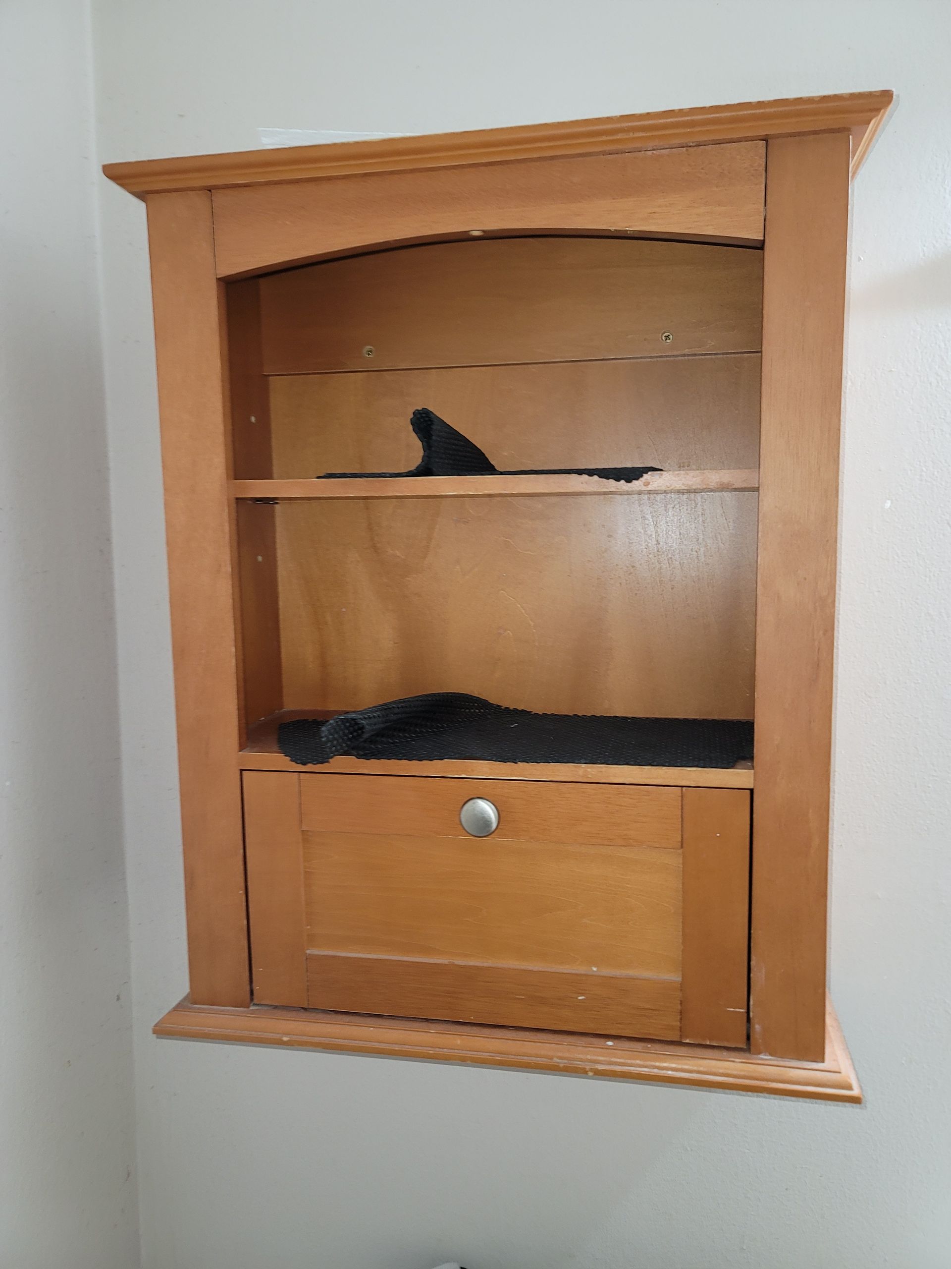 Wooden wall cabinet with shelves and a drawer, holding a few small dark objects.