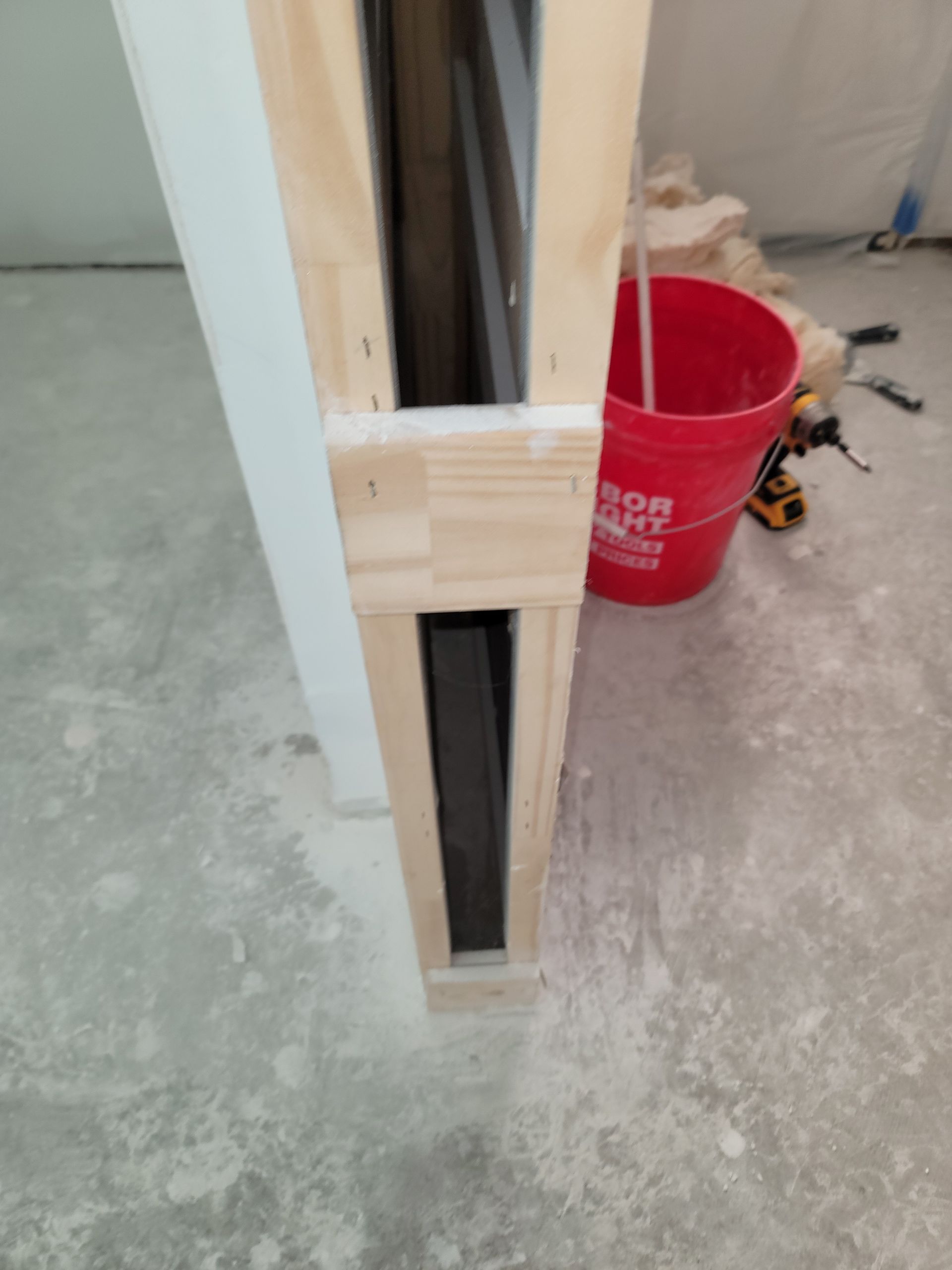 Wood-framed wall opening with exposed drywall edges beside a red bucket on a concrete floor.