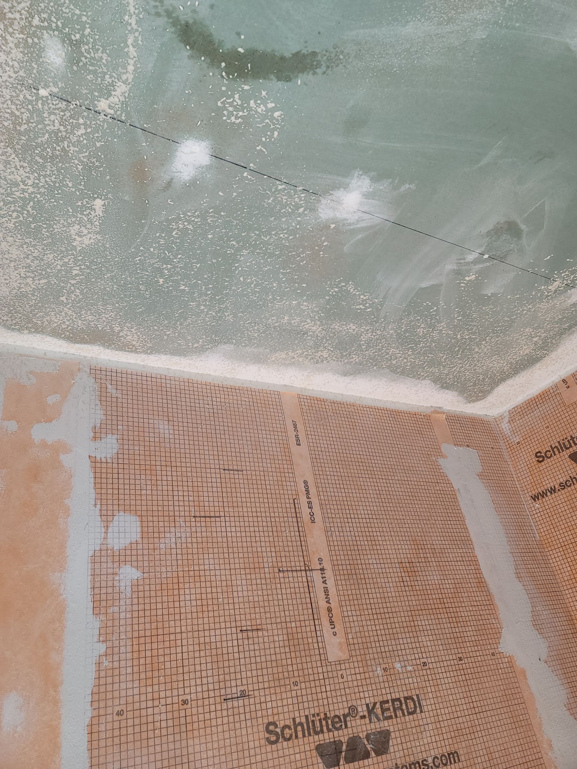 Partial view of a tan doormat on a tiled floor beside a green wall with peeling paint and white splatter marks