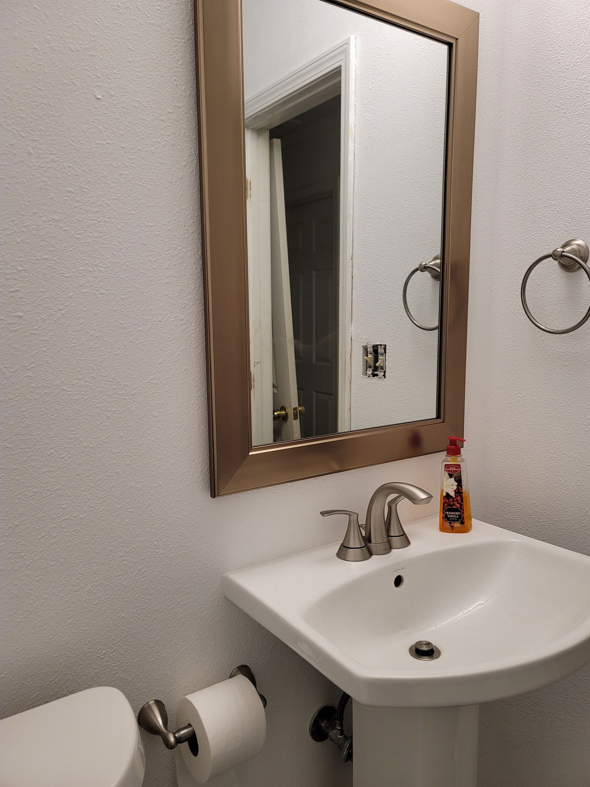 Small bathroom with white sink, mirror, toilet paper holder, and a decorative bottle on the sink counter.