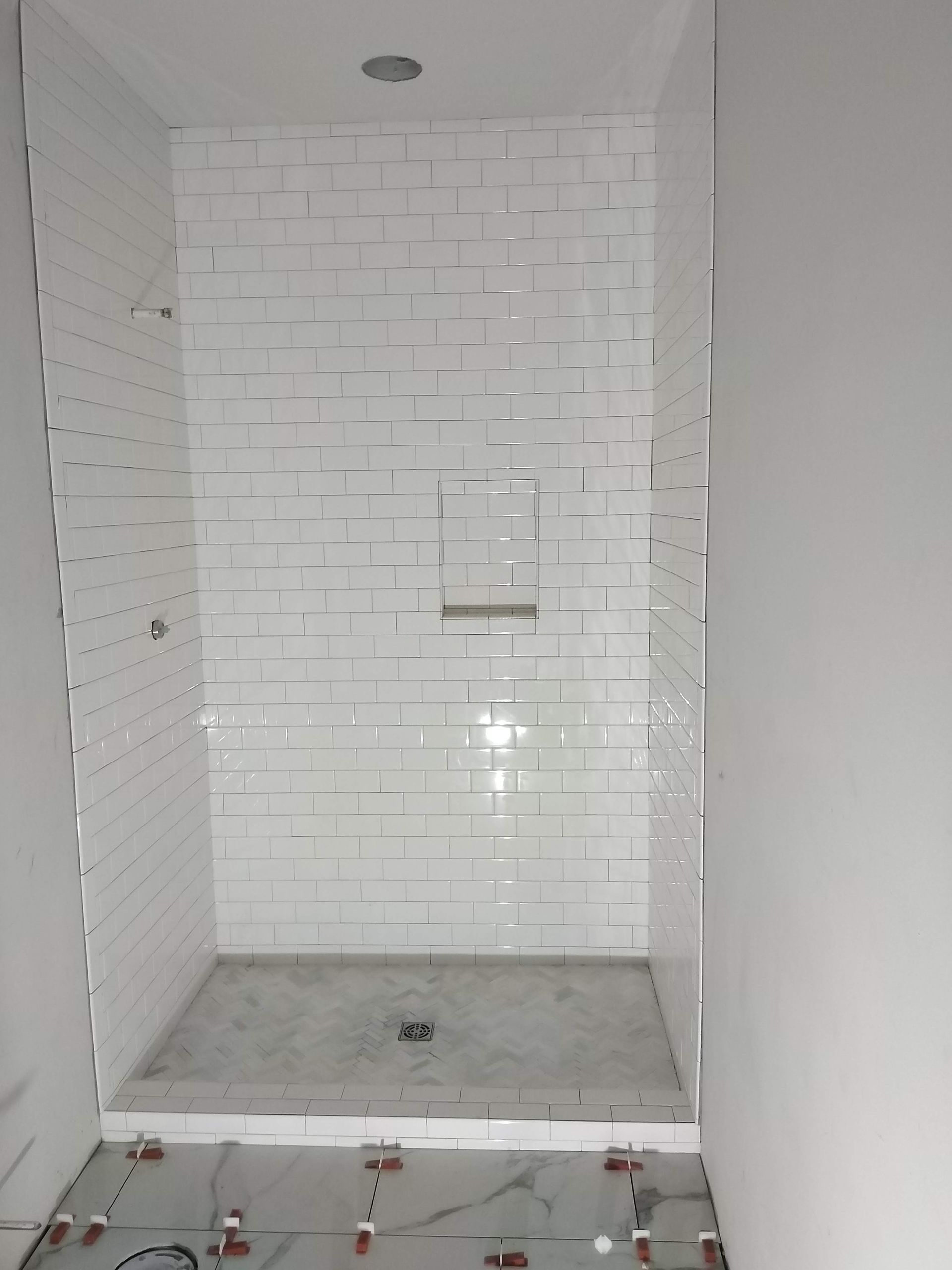 Small white-tiled shower stall under construction with unfinished floor and wall niche.