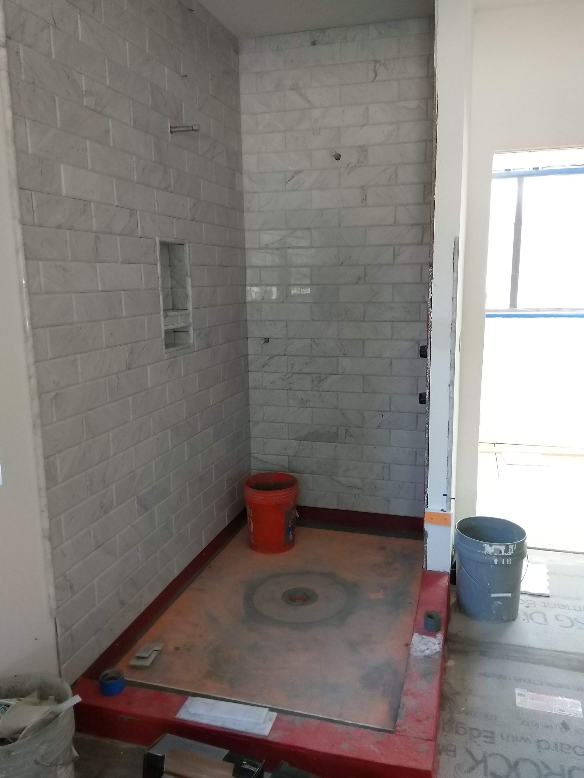 Small tiled shower stall under construction with a red bucket and blue bucket on the floor