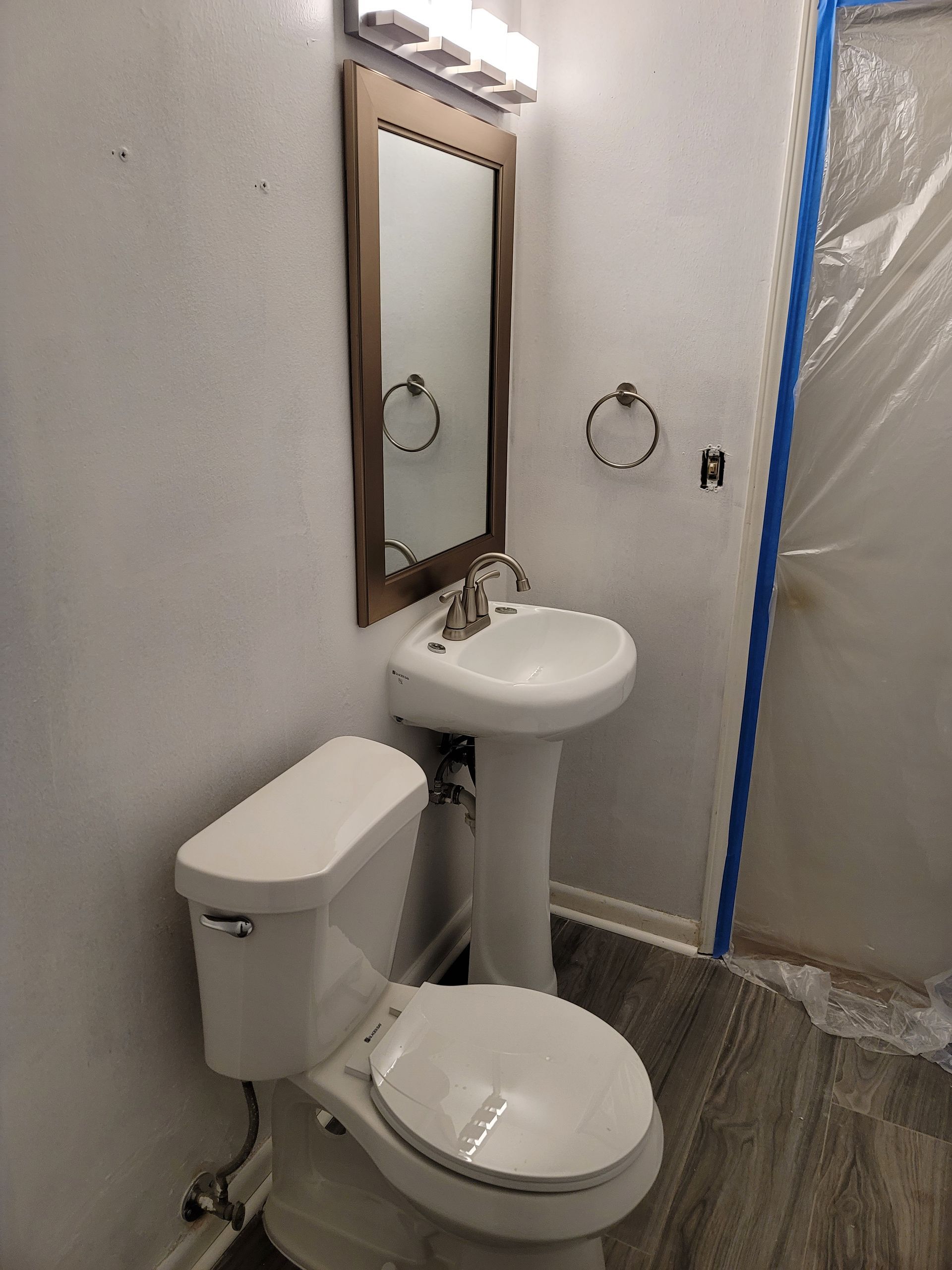 Small bathroom with toilet, pedestal sink, mirror, and shower area taped off with blue painter’s tape.