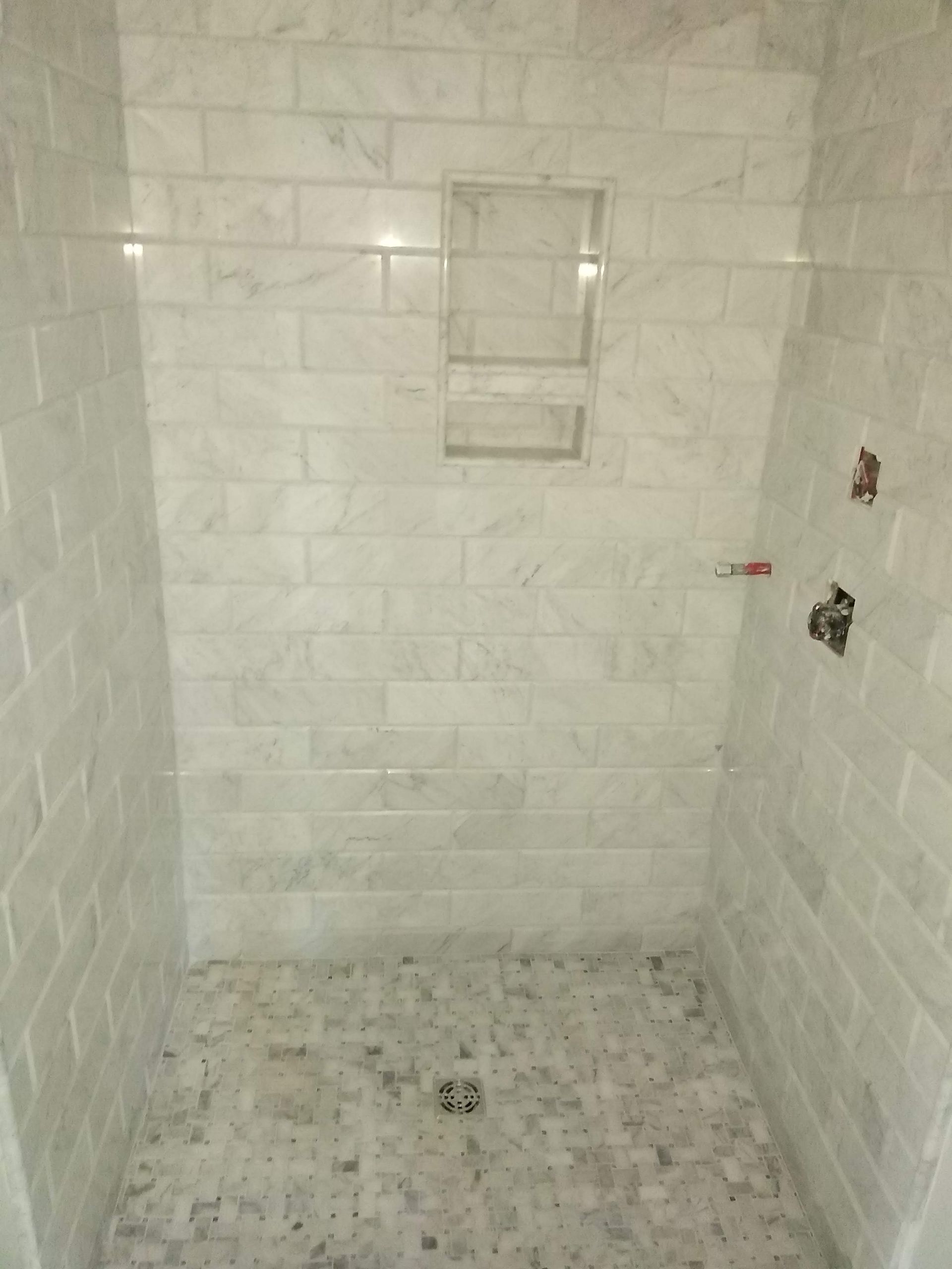 Empty tiled shower stall with a small recessed shelf and exposed plumbing on the right wall.