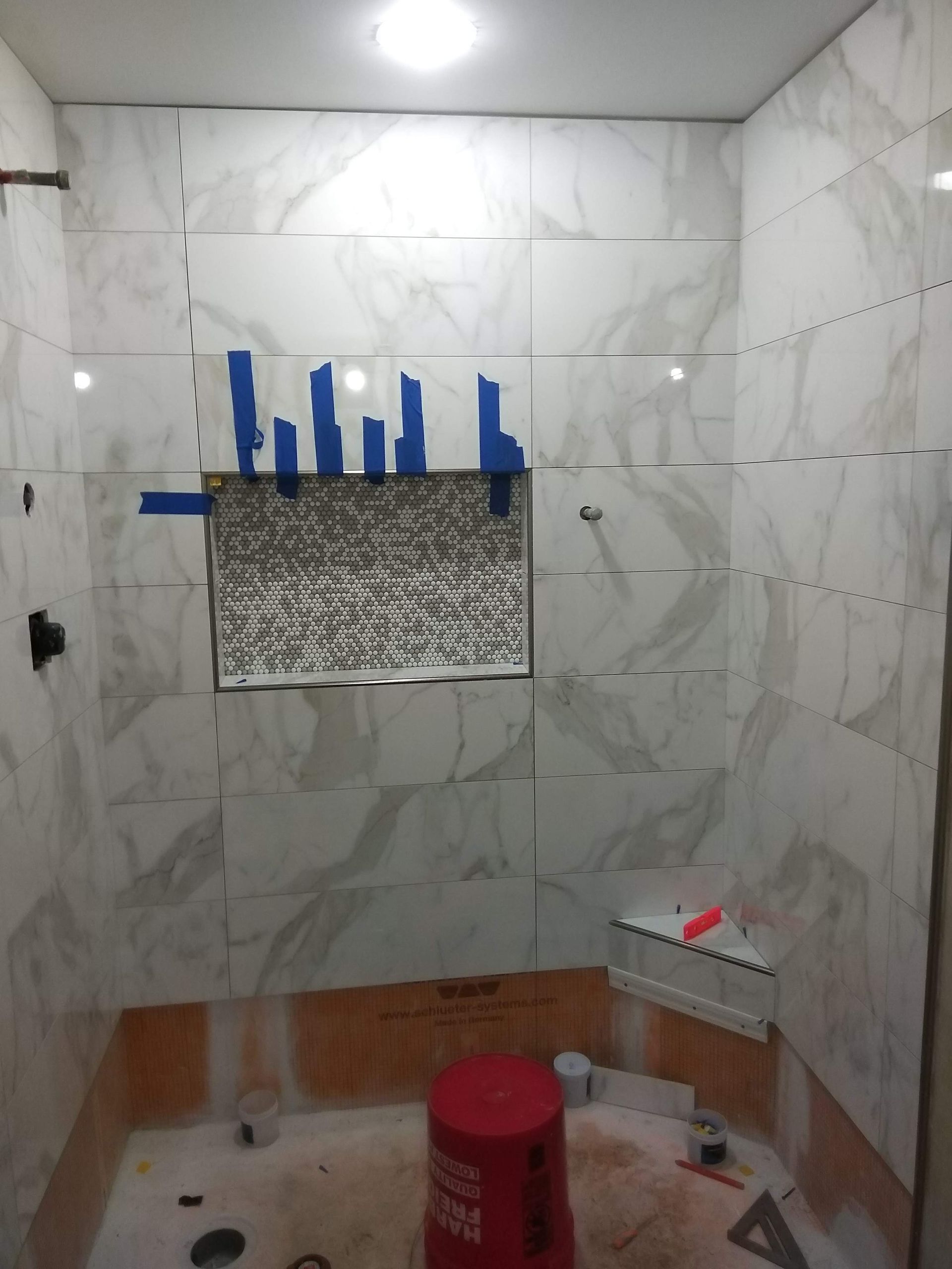 Partially tiled bathroom under renovation with a wall niche, tools, and a red bucket on the floor