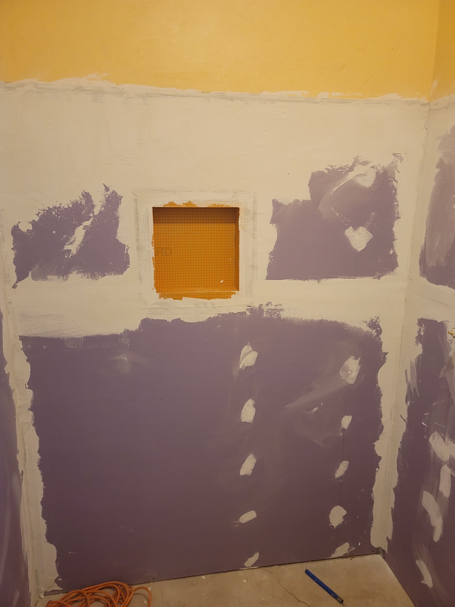 Partially patched drywall with purple joint compound around a small window opening on a beige wall.