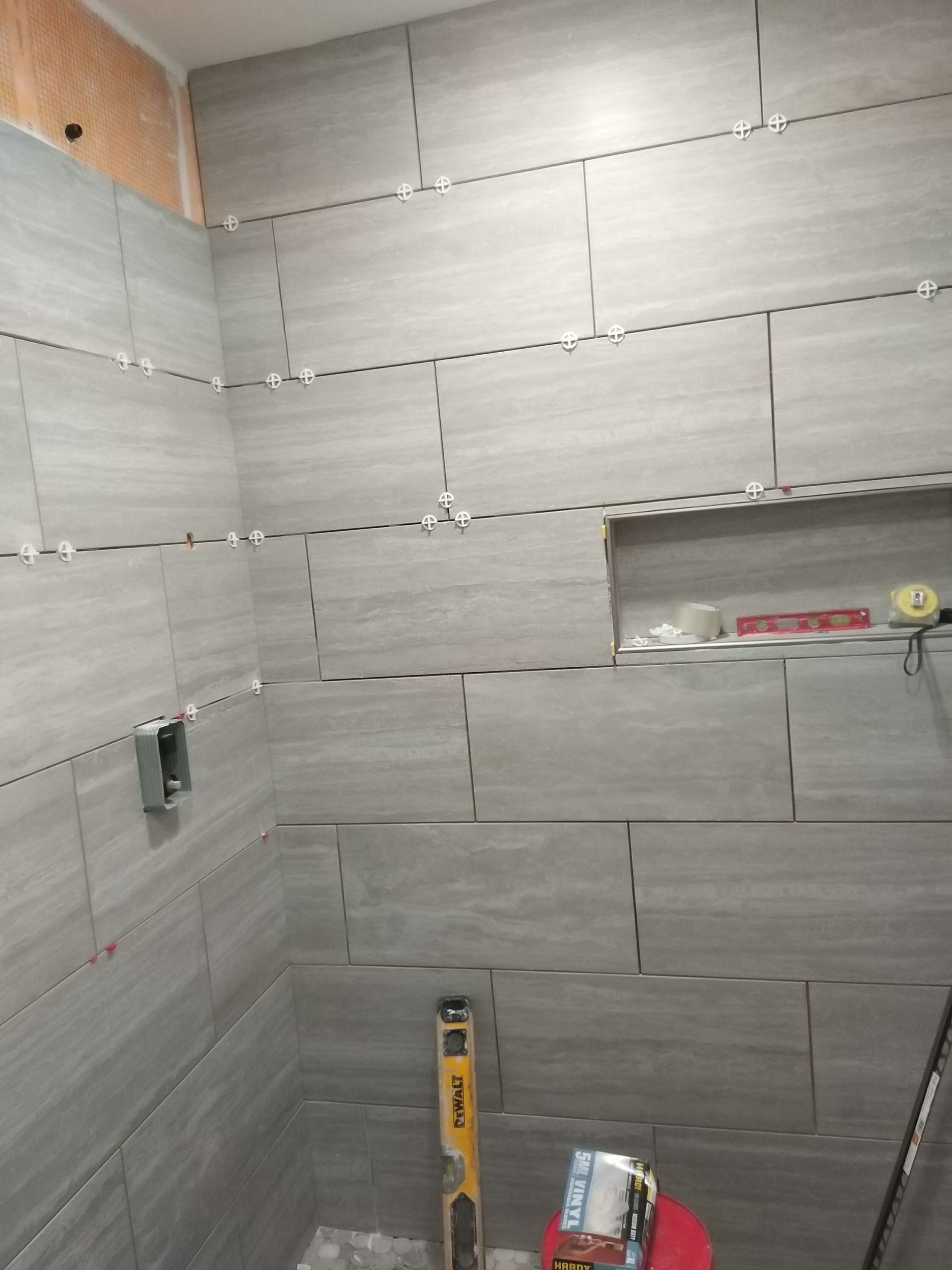 Partially tiled bathroom shower wall with gray tiles, exposed seams, and construction tools on the floor