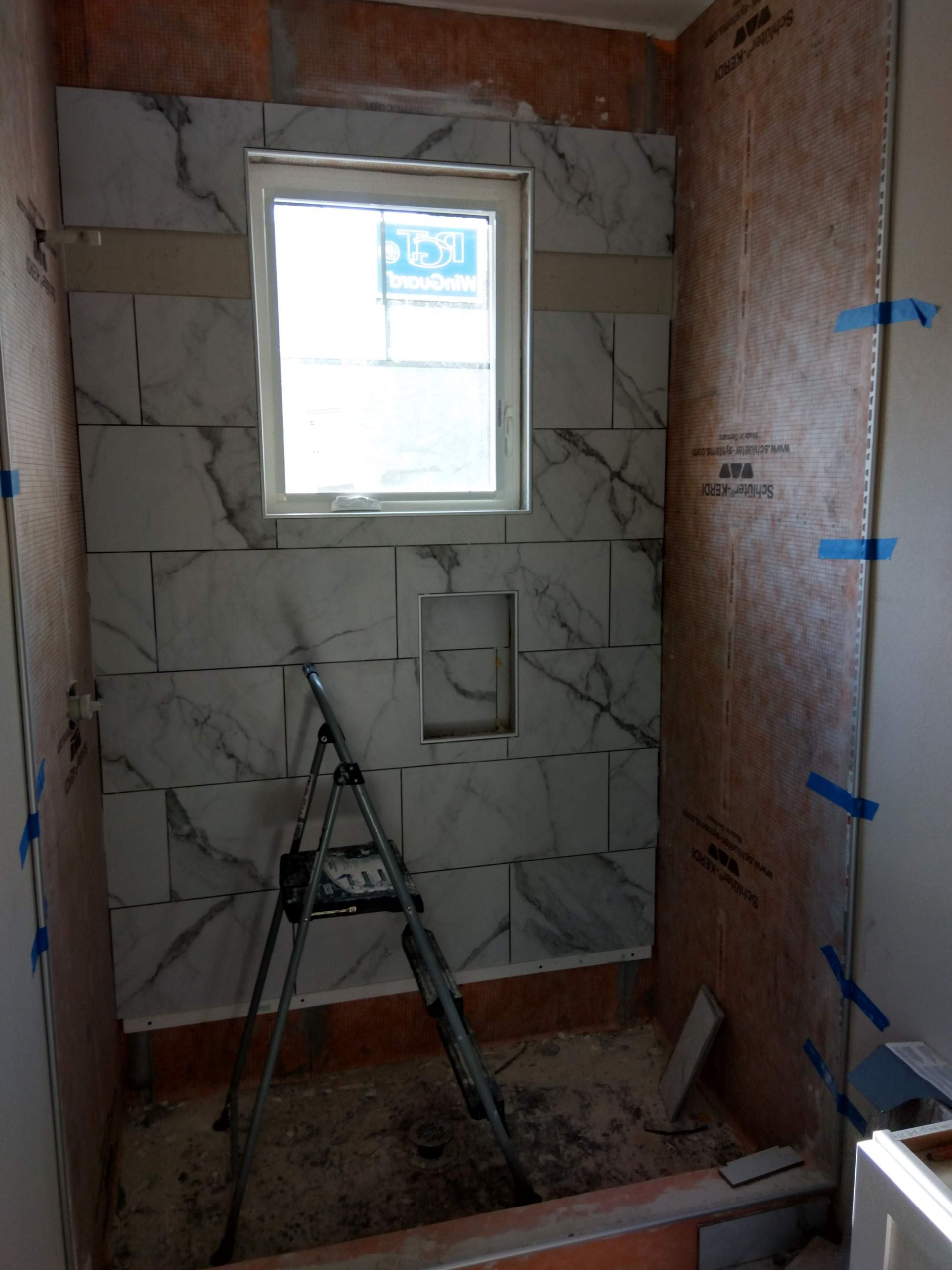 Small bathroom under construction with tiled walls, a window, and a ladder on the floor.