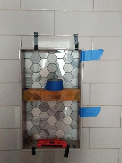 Wall-mounted metal shelf with hex tile backing, blue tape, and a wooden shelf on a tiled wall.