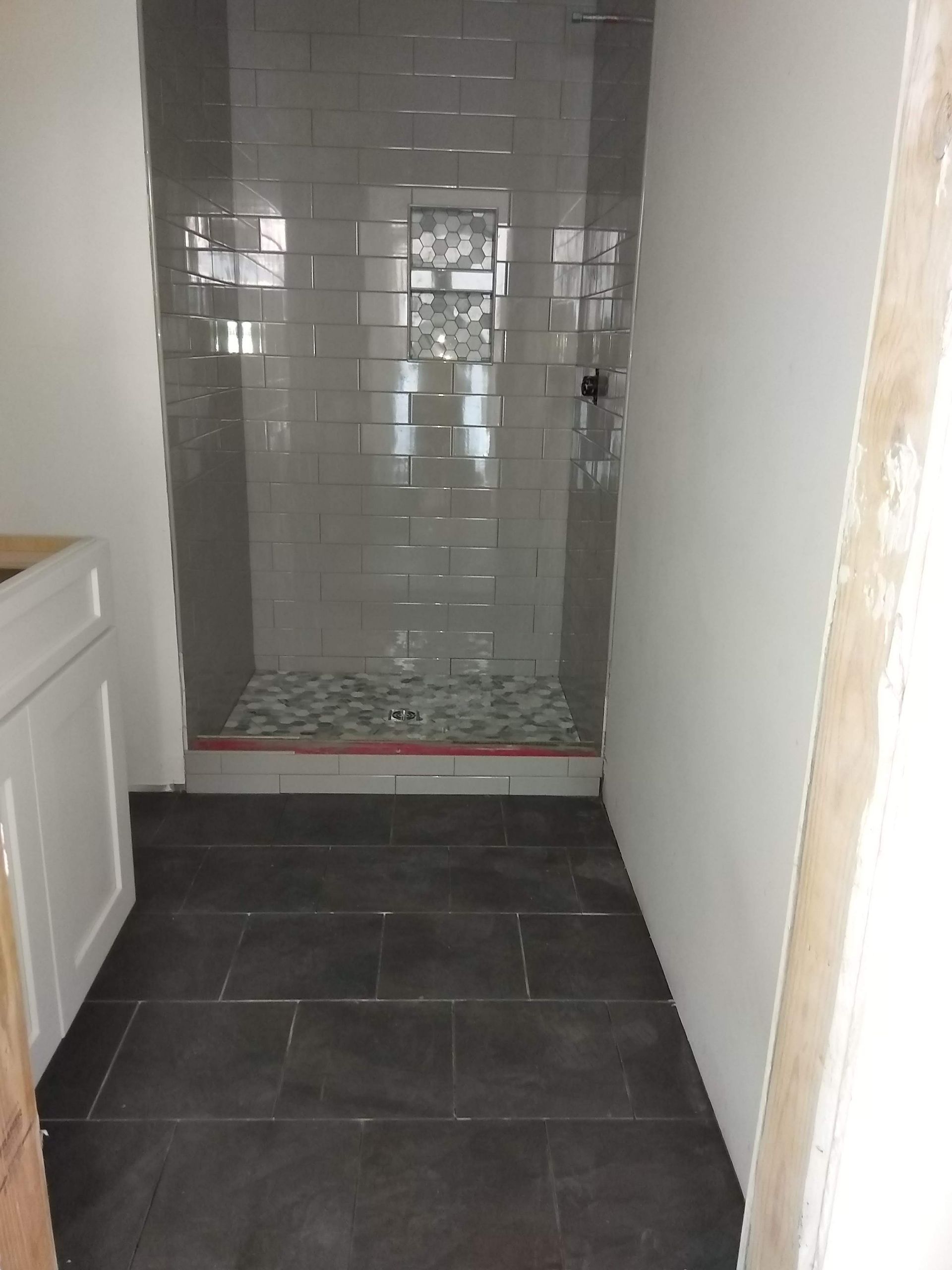 Narrow tiled bathroom with a walk-in shower at the far end and dark floor tiles in the foreground