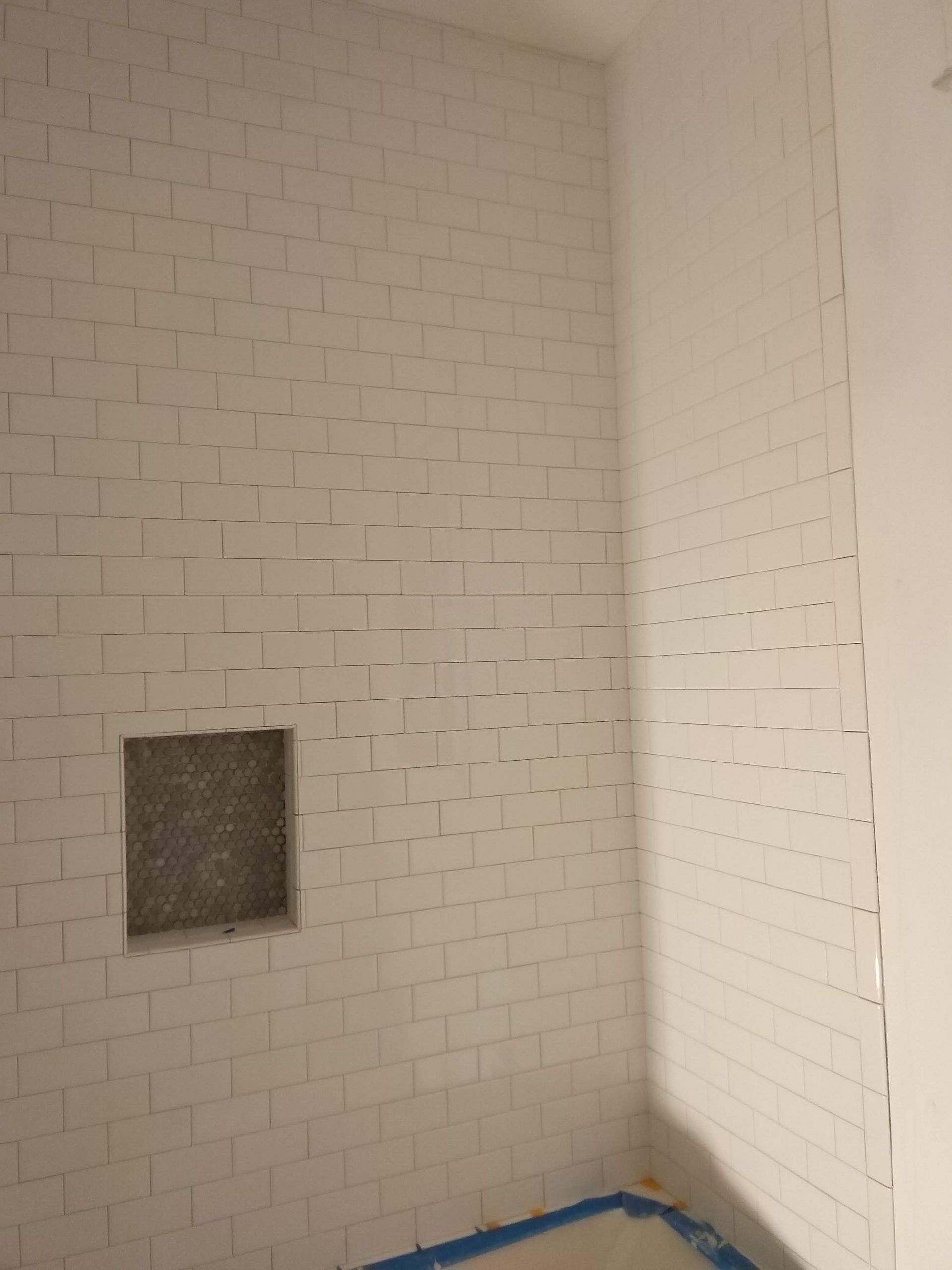 Empty tiled shower corner with a small dark recessed opening and blue tape along the floor edge