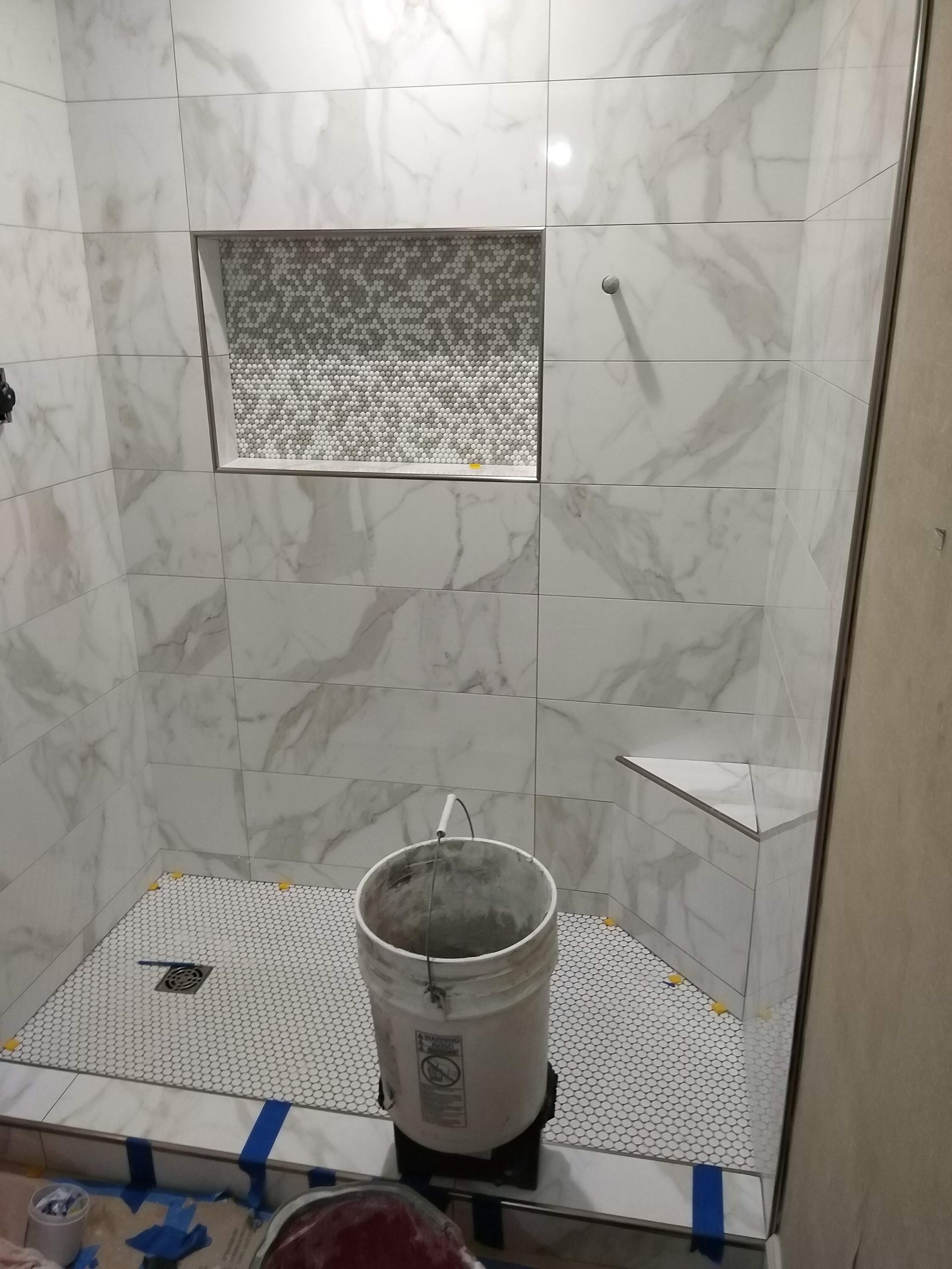 Tiled shower under renovation with a bucket on the floor and a recessed wall niche.