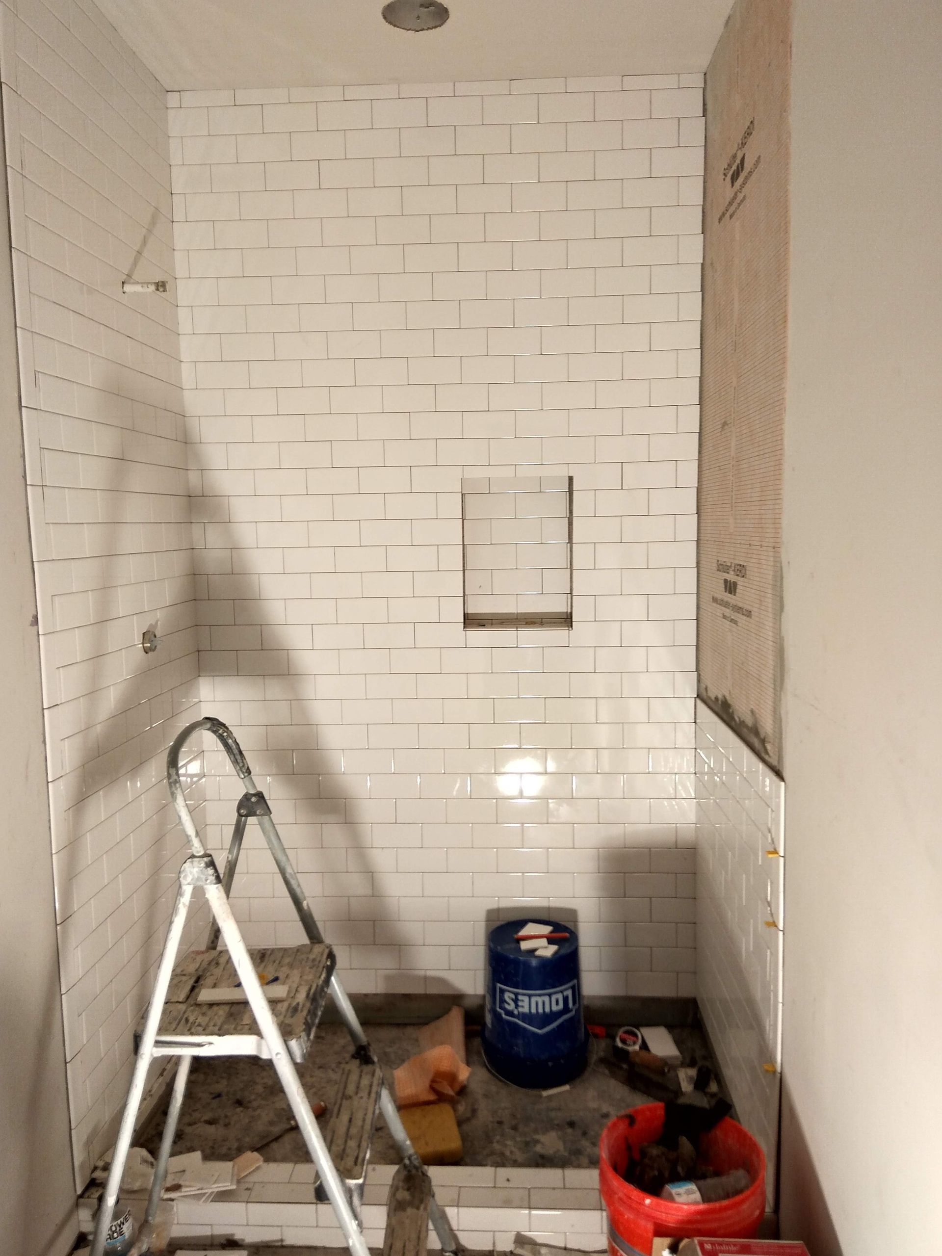 Small bathroom under renovation with tiled walls, step ladder, paint can, and tools on the floor.